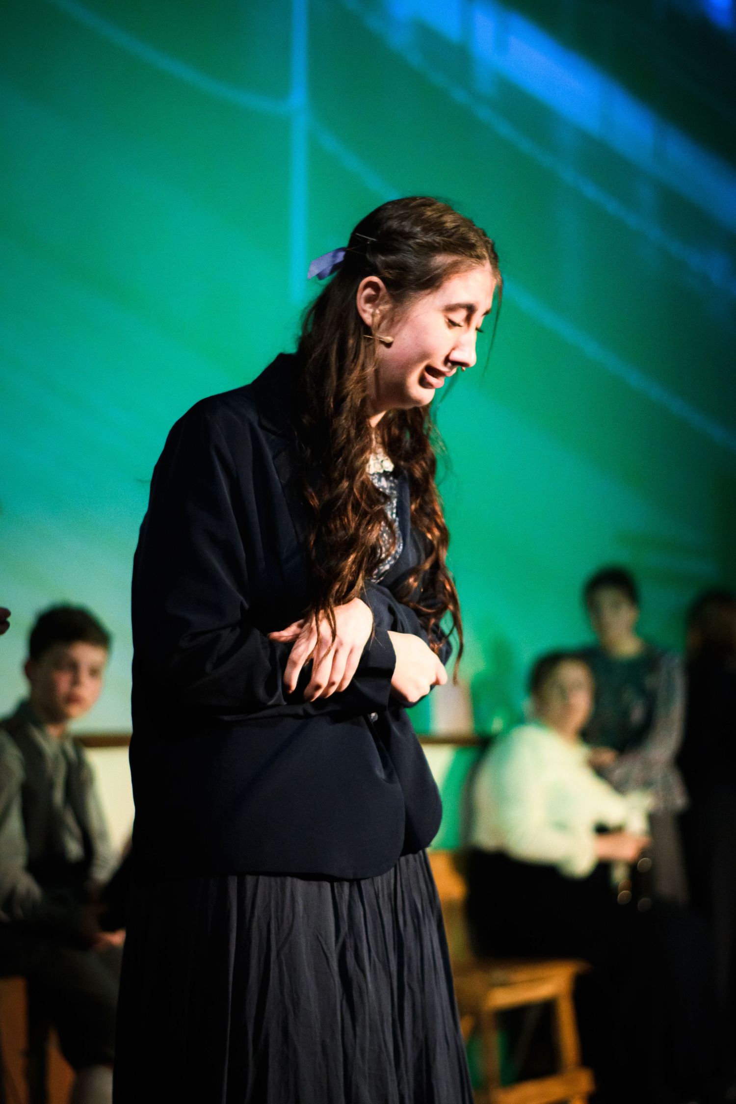 A performer in a dark dress laughs during a theatrical production with green lighting effects in the background.
