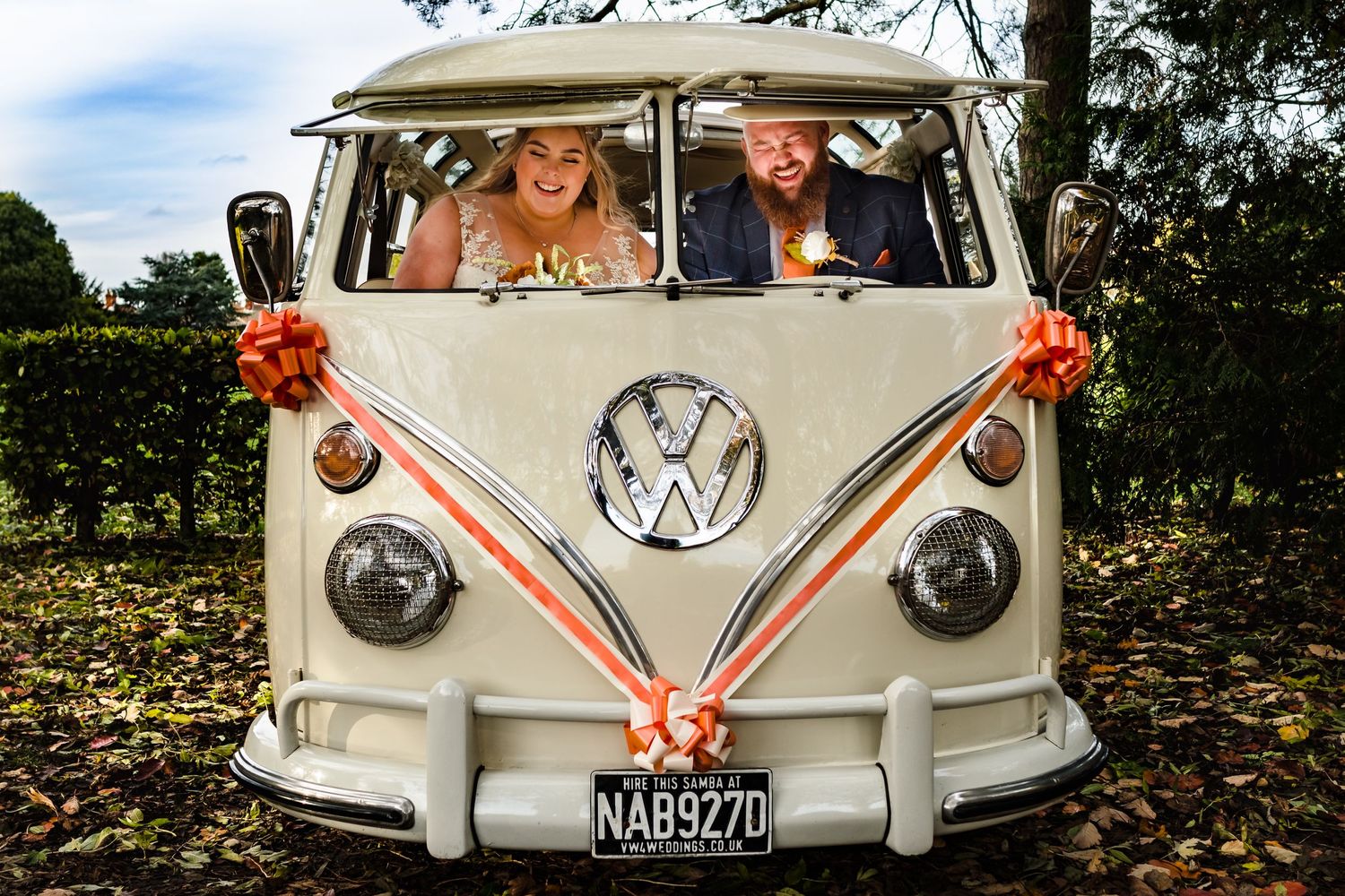 A happy couple poses in a vintage white Volkswagen bus decorated with orange flowers for their wedding.