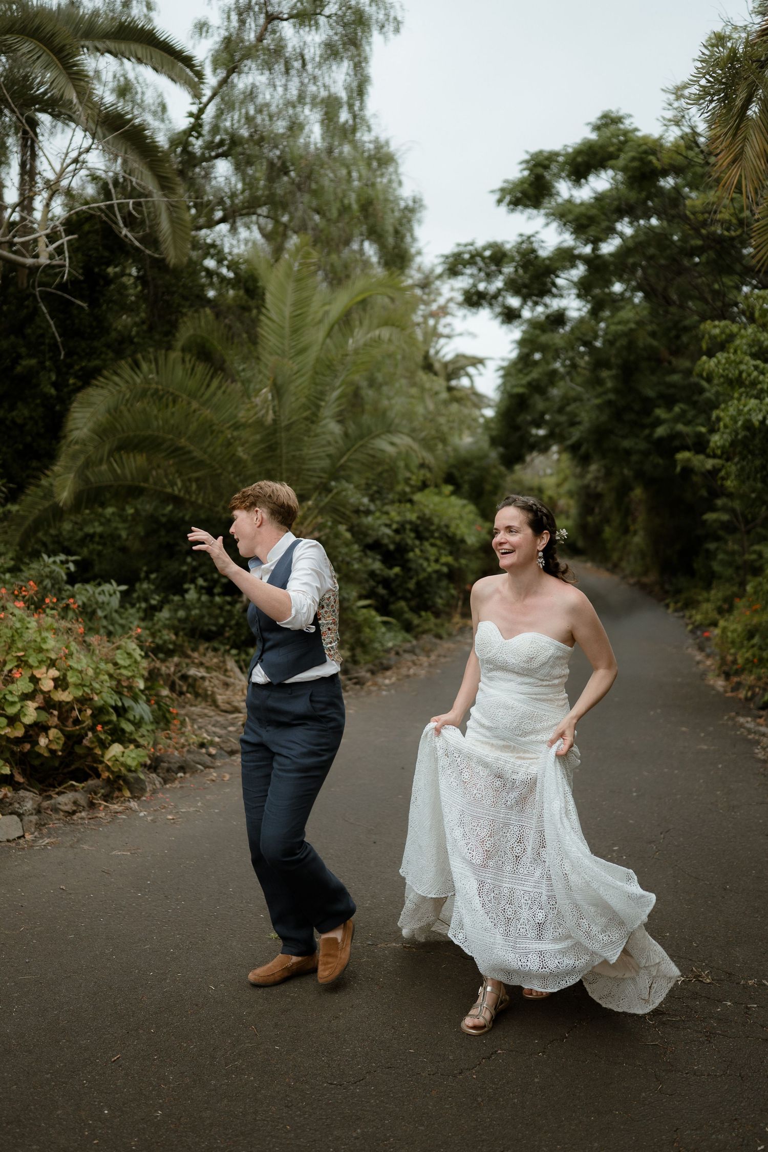 Joyful couple in wedding attire dancing on a tree-lined path, bride in white lace gown lifting her dress.