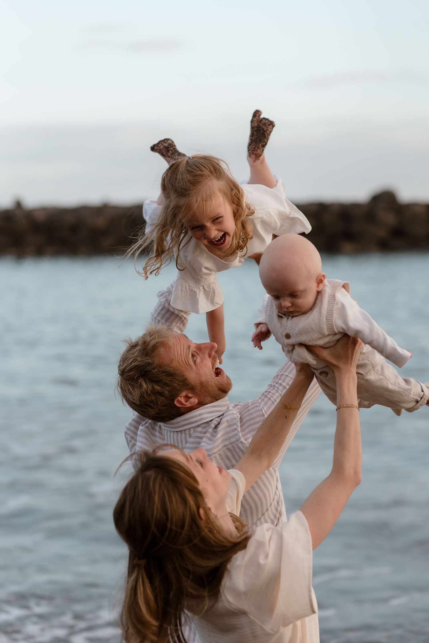 Family of three enjoying a beach day, parents lifting laughing toddler and baby in the air near ocean.