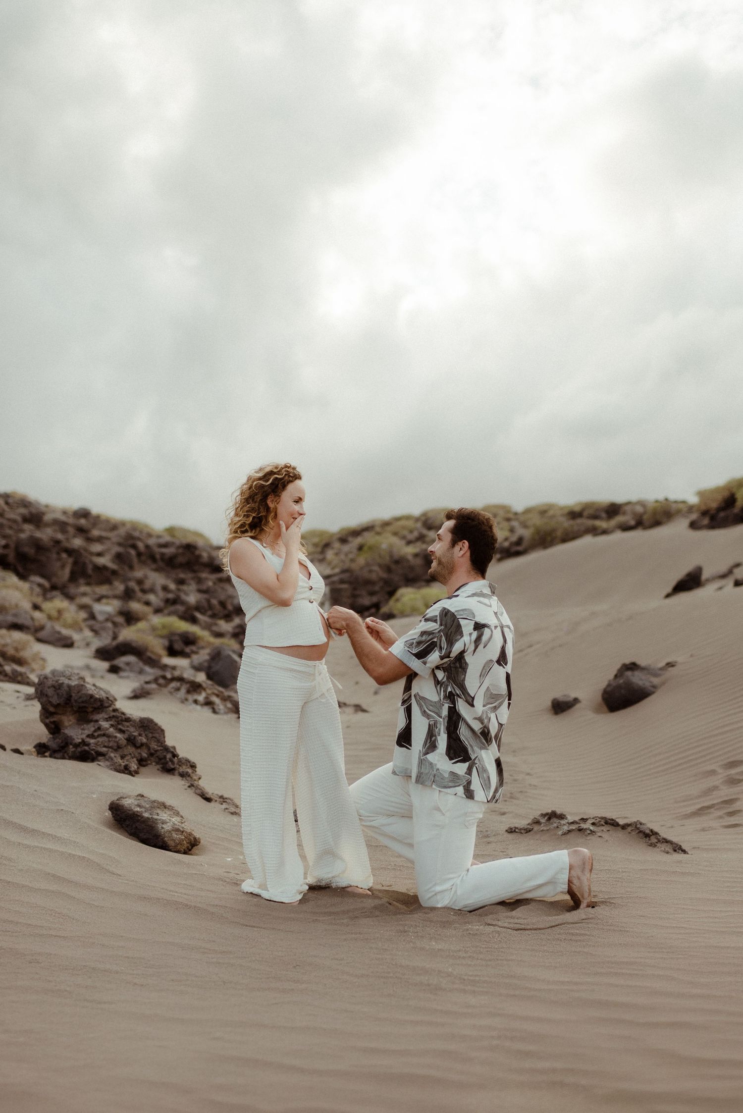 Two people share an intimate moment on sandy beach dunes against cloudy sky.
