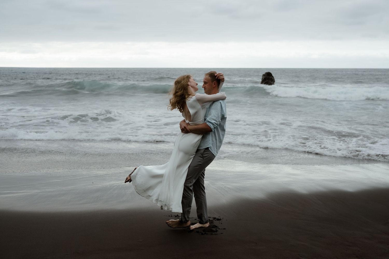 Couple sharing a romantic embrace on a black sand beach with ocean waves, bride in white dress, groom in gray suit.