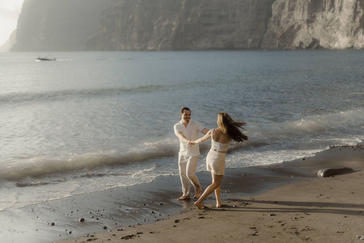 A couple in white outfits dances joyfully on a beach with dramatic cliffs in the background.