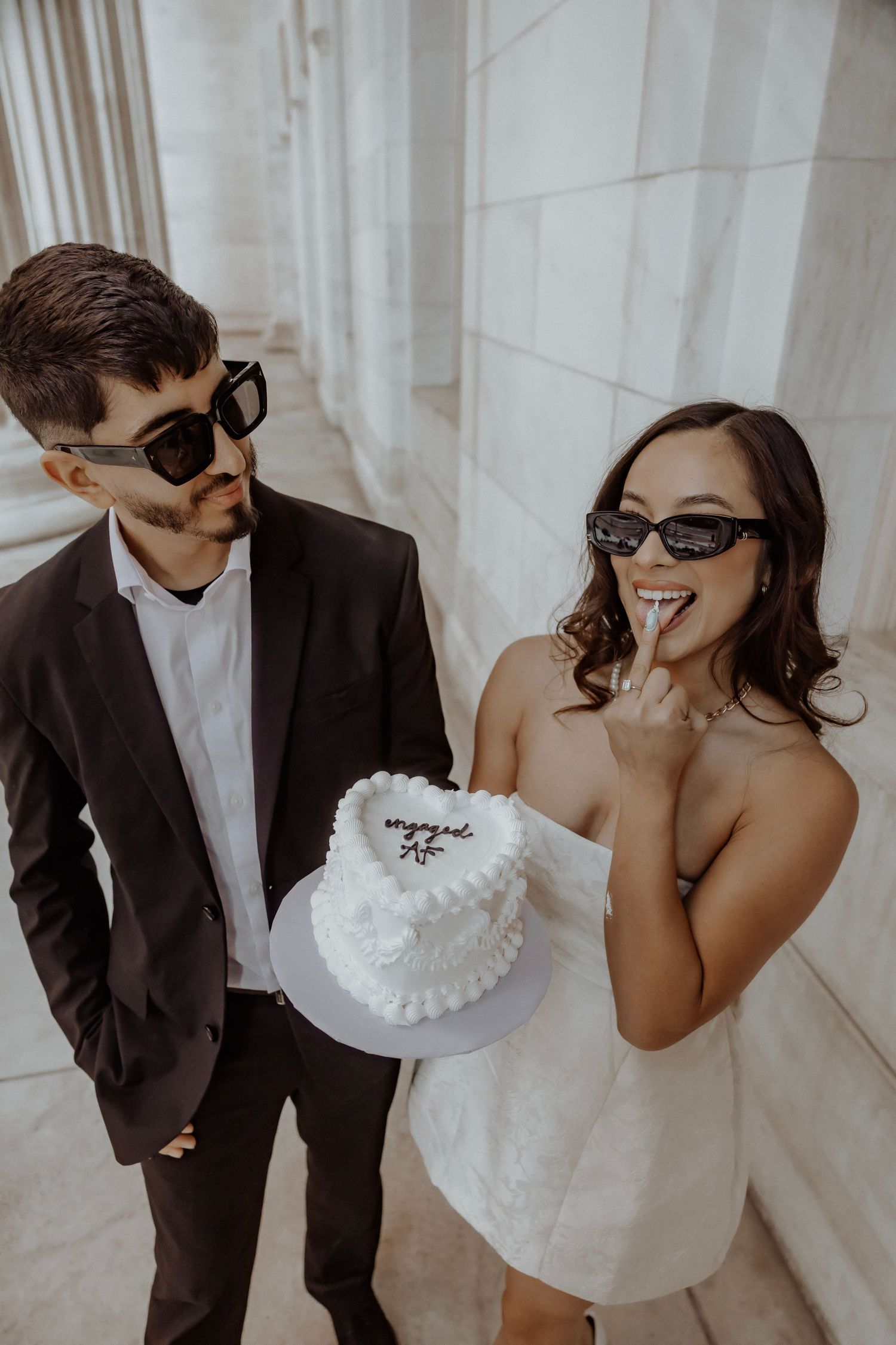 Engaged couple, woman is holding a heart shape cake licking ring finger showing off engagement ring