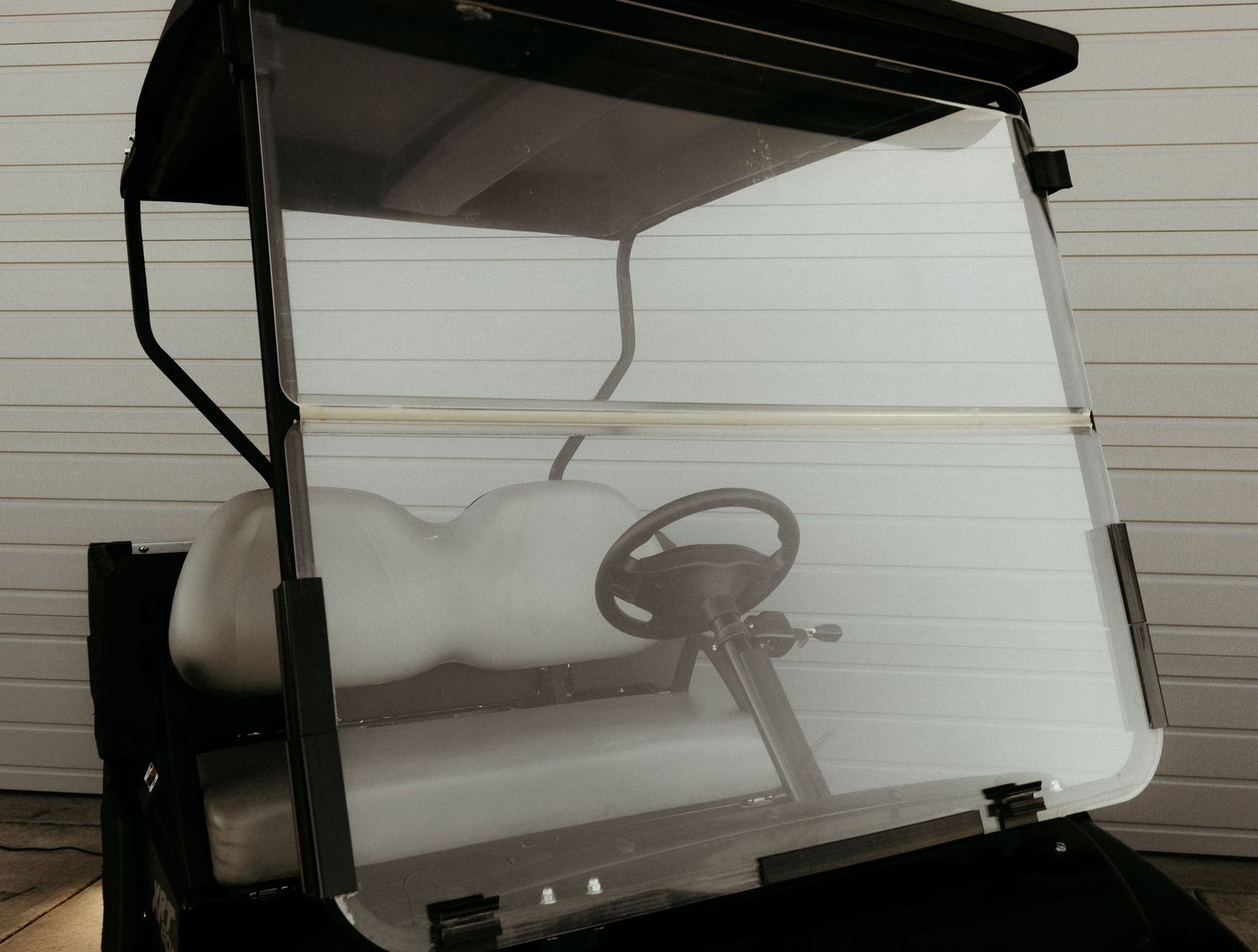 Clear Club Car golf cart windshield with dark canopy top mounted against white siding background.