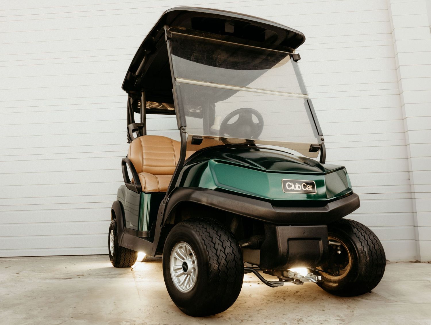 Teal Club Car golf cart with tan seats and black canopy parked against a white wall.