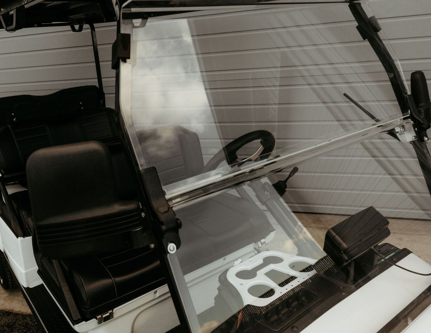 Close up of a golf cart windshield and steering wheel with black seats visible against a white garage door.