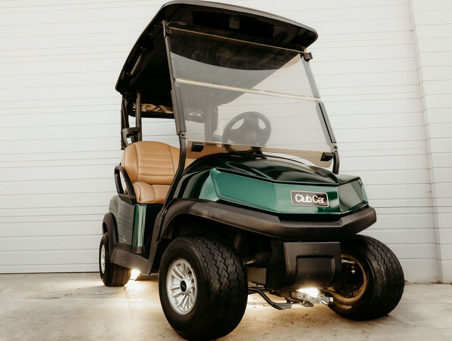 Club Car Precedent golf cart in dark green with tan seats parked against a white garage wall.