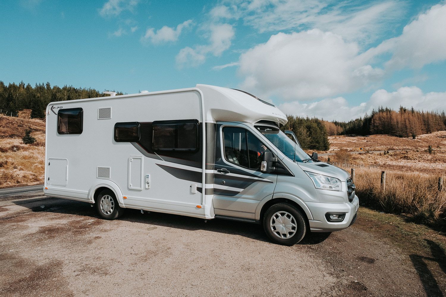 White motorhome camper van parked on a dirt road with scenic mountains and forest in the background.
