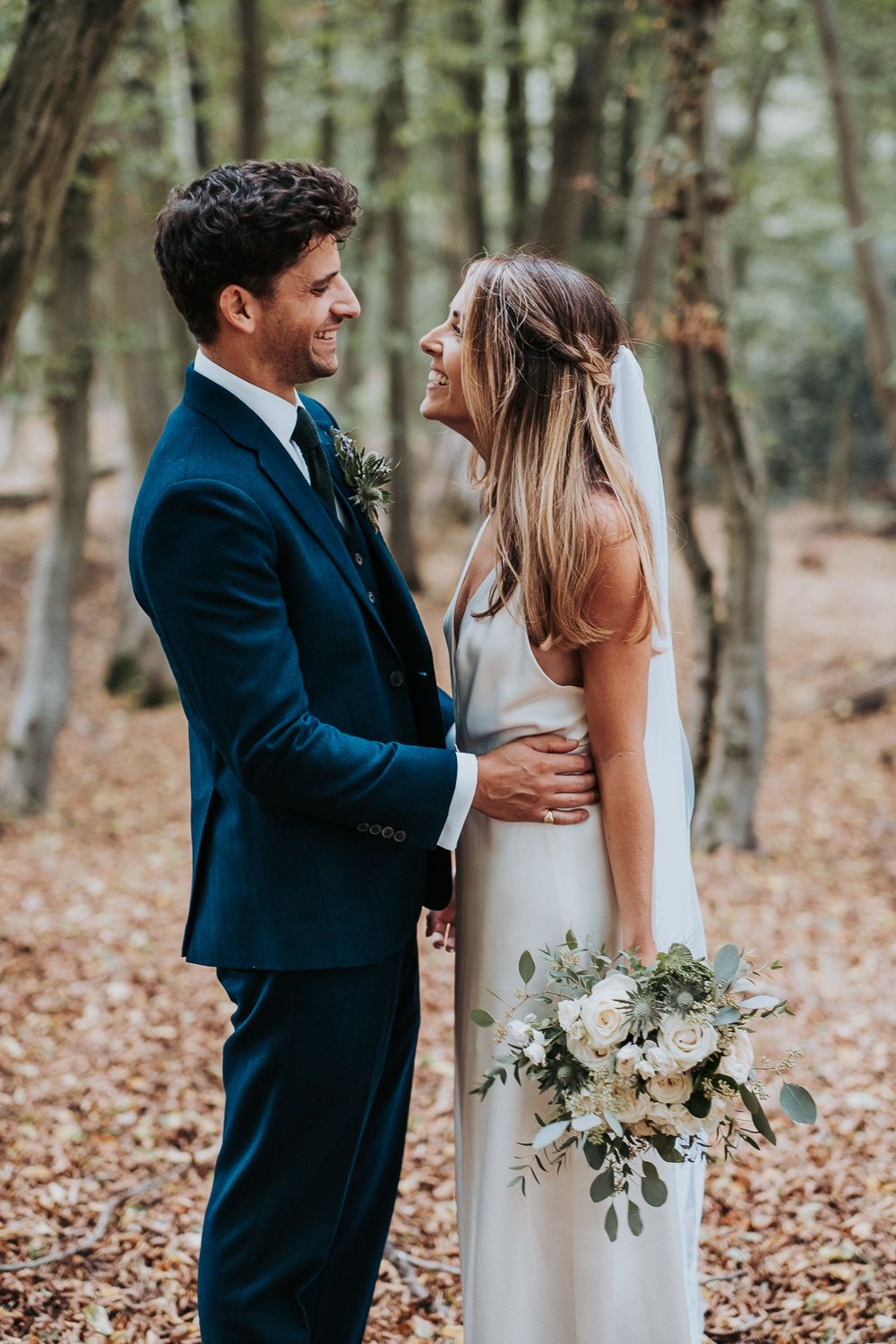 An intimate wedding portrait shows a couple gazing at each other in a wooded setting with autumn leaves.