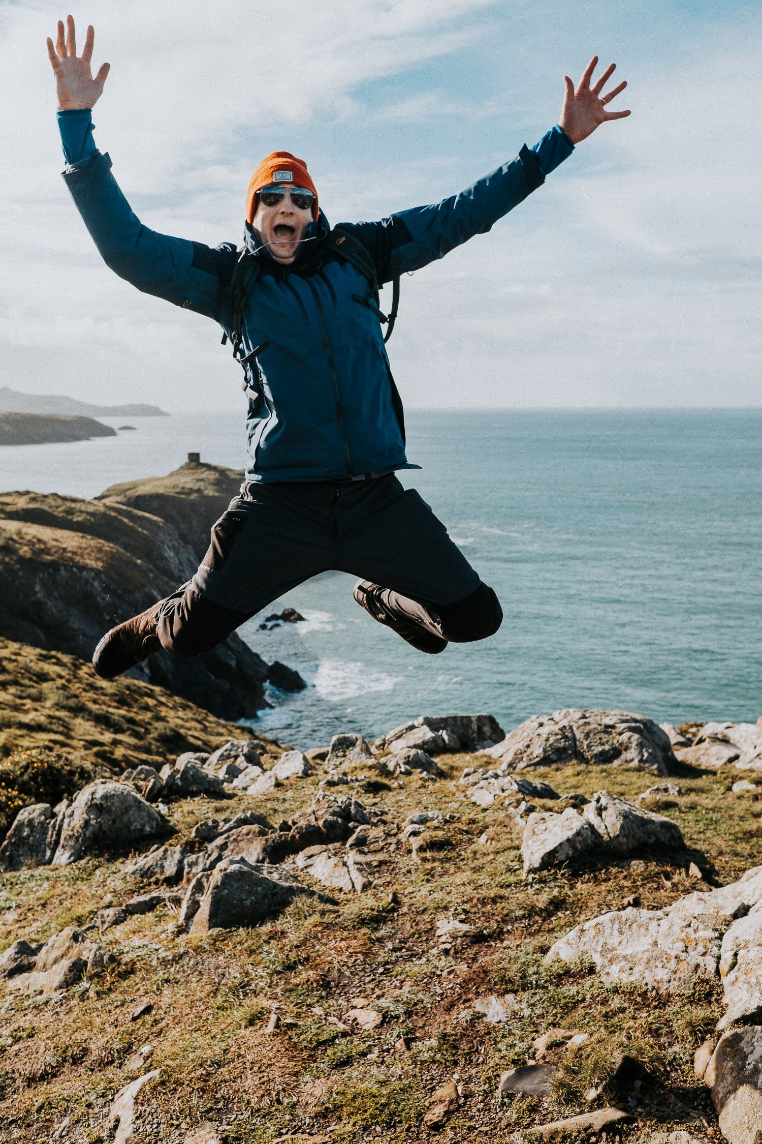 Adventurous person jumping with arms raised on rocky coastal cliff overlooking the ocean.