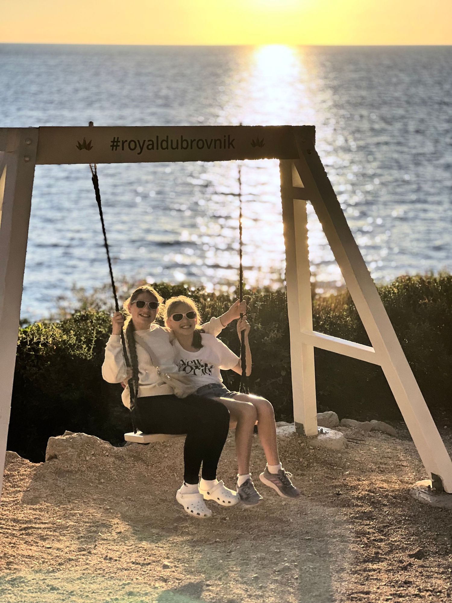 Two people enjoying sunset on a wooden swing marked with #royaldubrovnik by the ocean.
