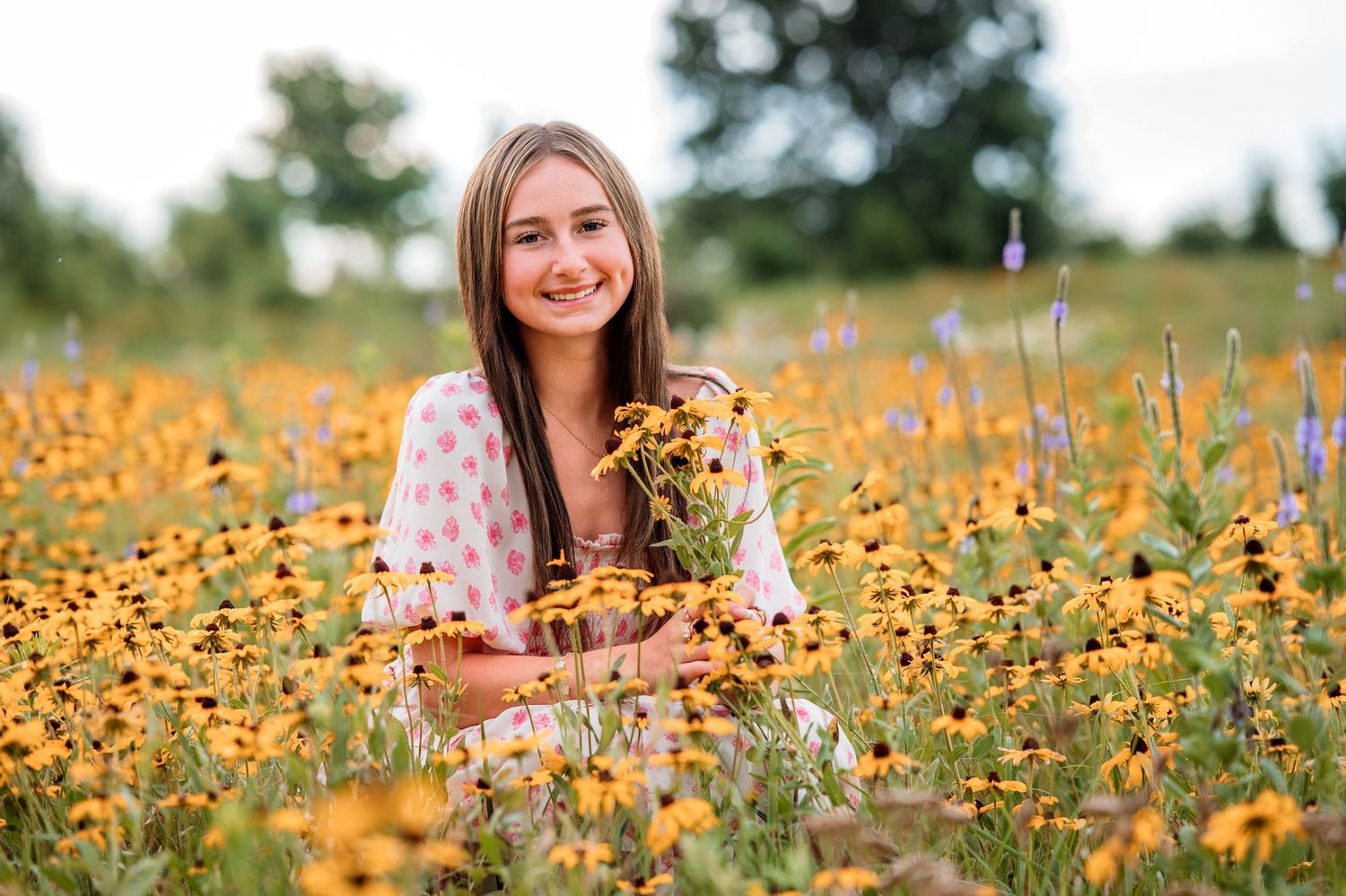 Smiling young woman holding yellow black-eyed Susan wildflowers while sitting in a colorful meadow field outdoors.
