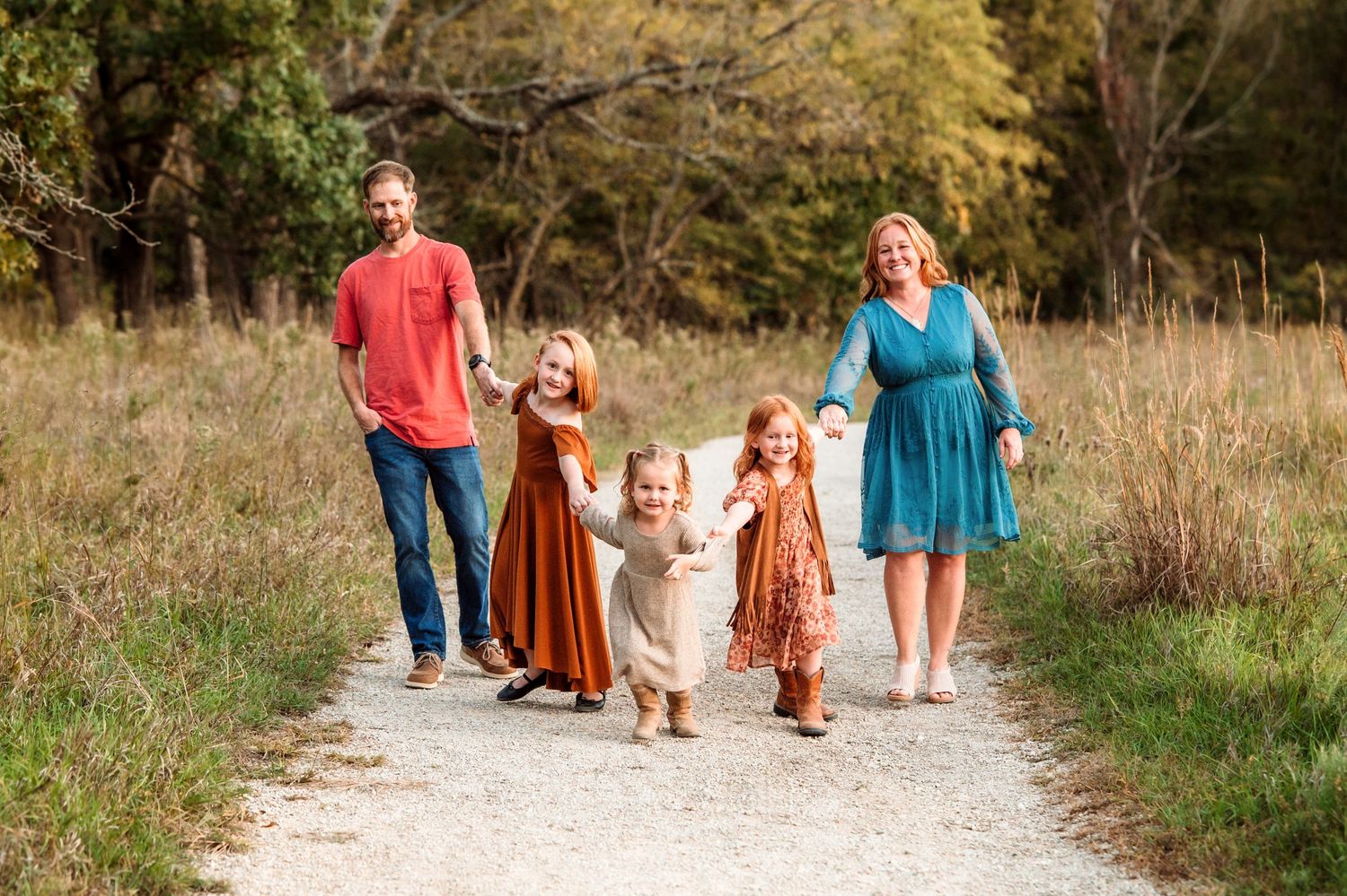 Family walking together on a dirt path through autumn fields with trees in the background.