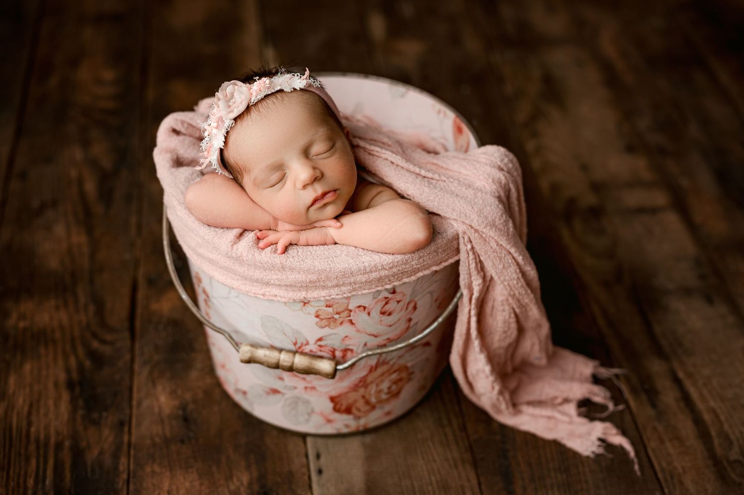 Sleeping newborn nestled in pink fabric inside a decorative round container on wooden surface.