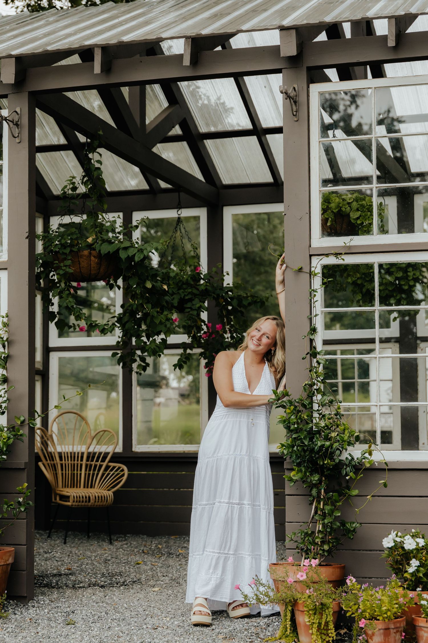 Woman in white maxi dress posing by a charming glass greenhouse with hanging plants and flowers.