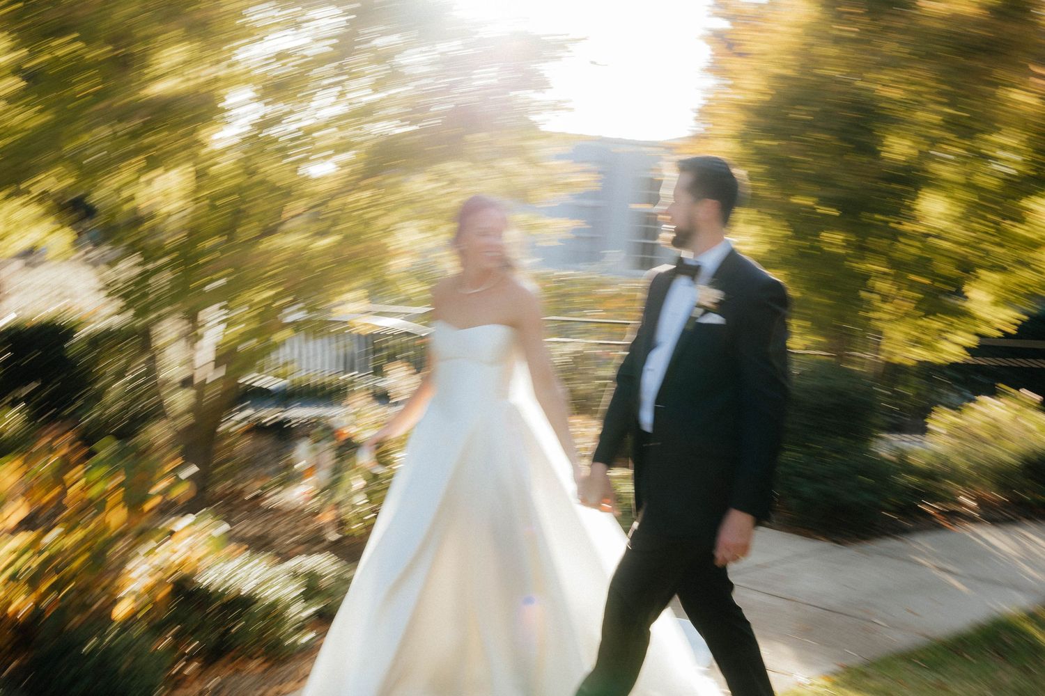 A couple in wedding attire strolls through a sunlit garden path with golden autumn foliage overhead.