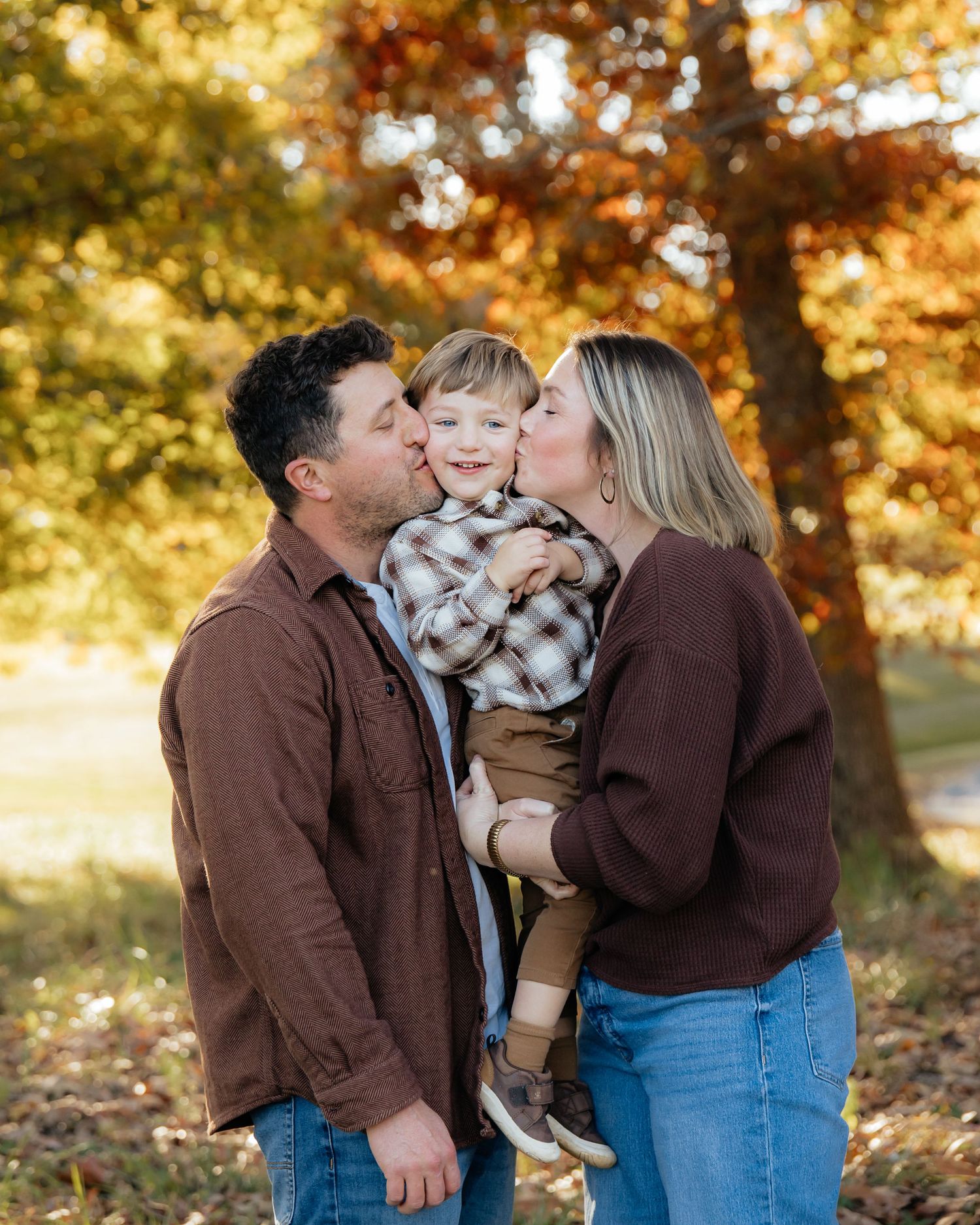 A family shares a tender moment together in a park during autumn with golden fall foliage in the background.