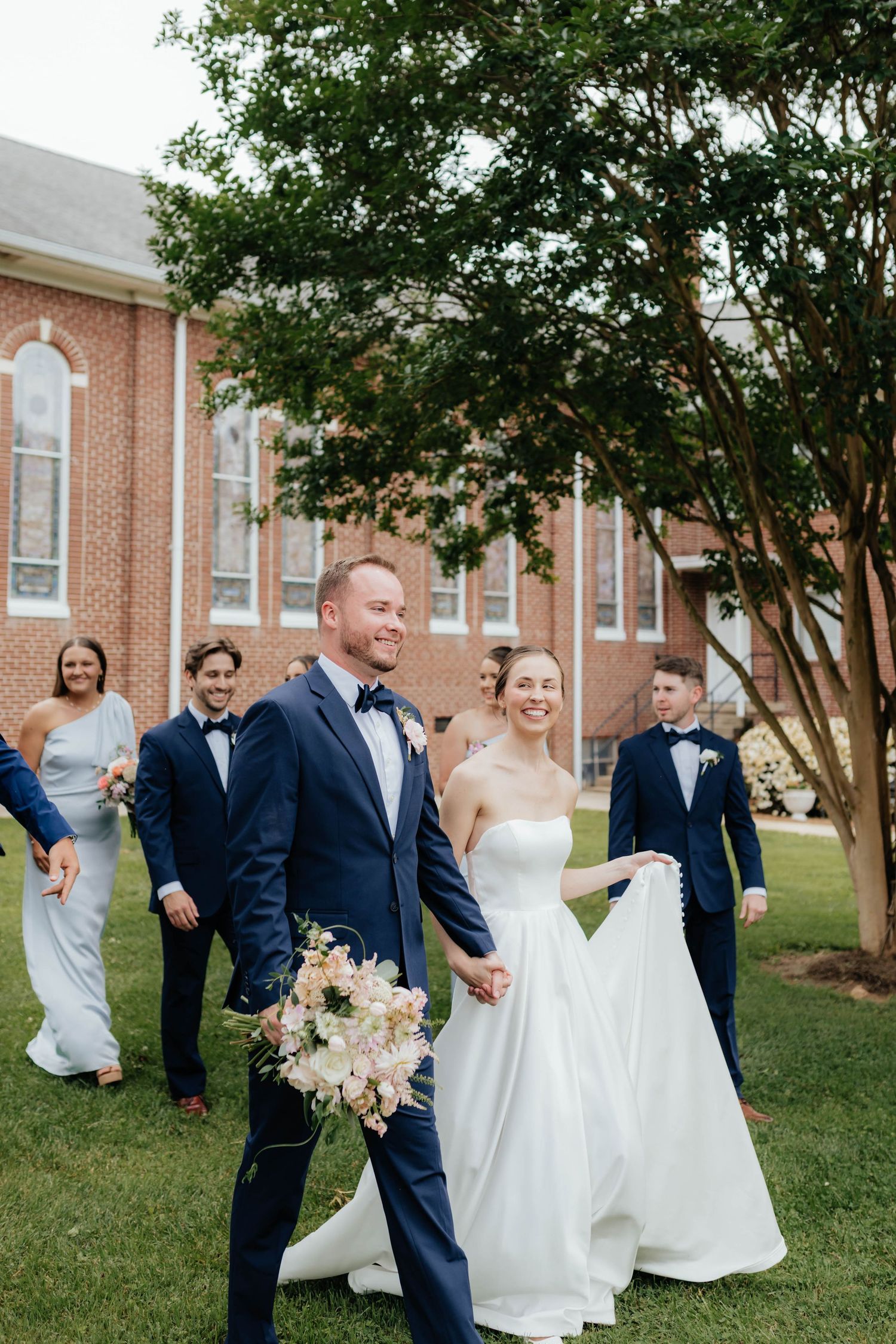 A joyful wedding party laughs together on a lawn in front of a brick church building.