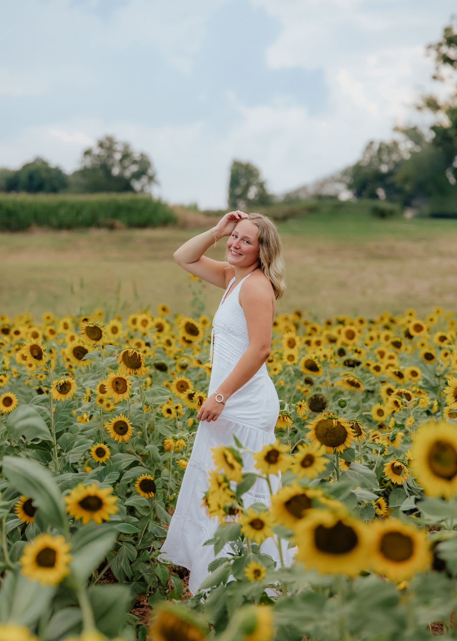 Smiling blonde woman in white dress standing in a vibrant sunflower field on a sunny day.
