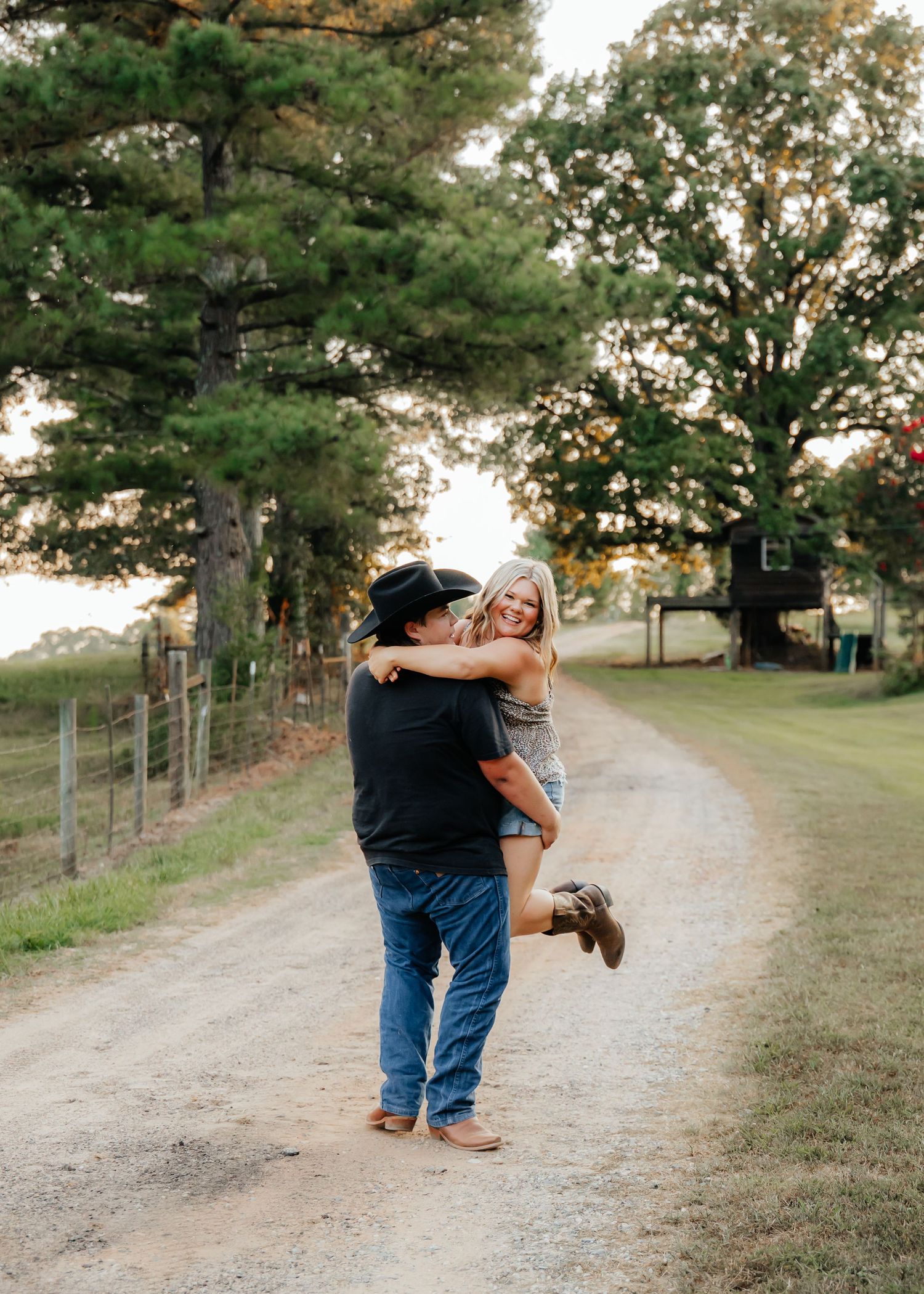 A couple shares a romantic dance sequence on a rustic dirt path surrounded by tall pine trees at sunset.