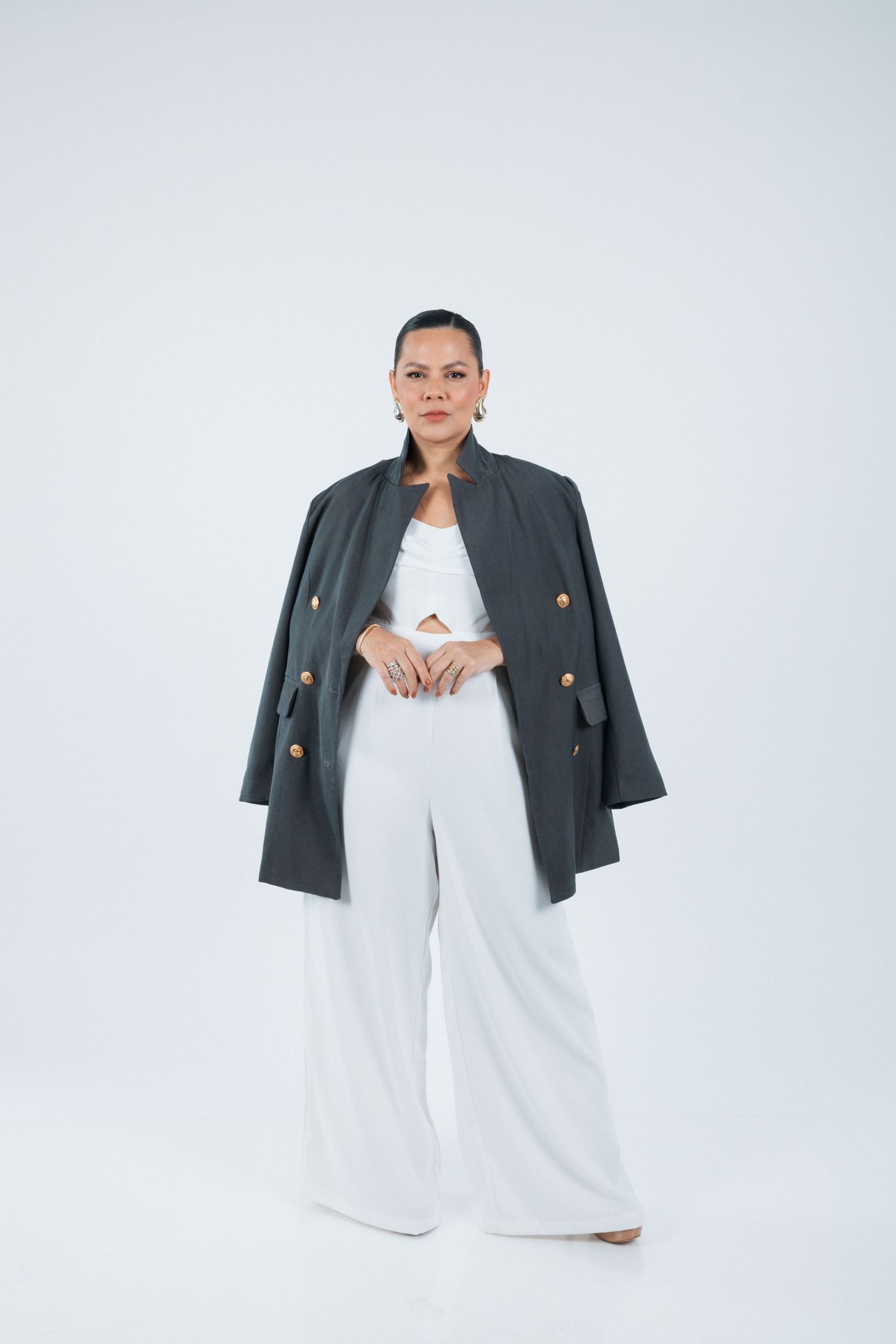 Professional model in white pants and grey blazer poses confidently against white background.
