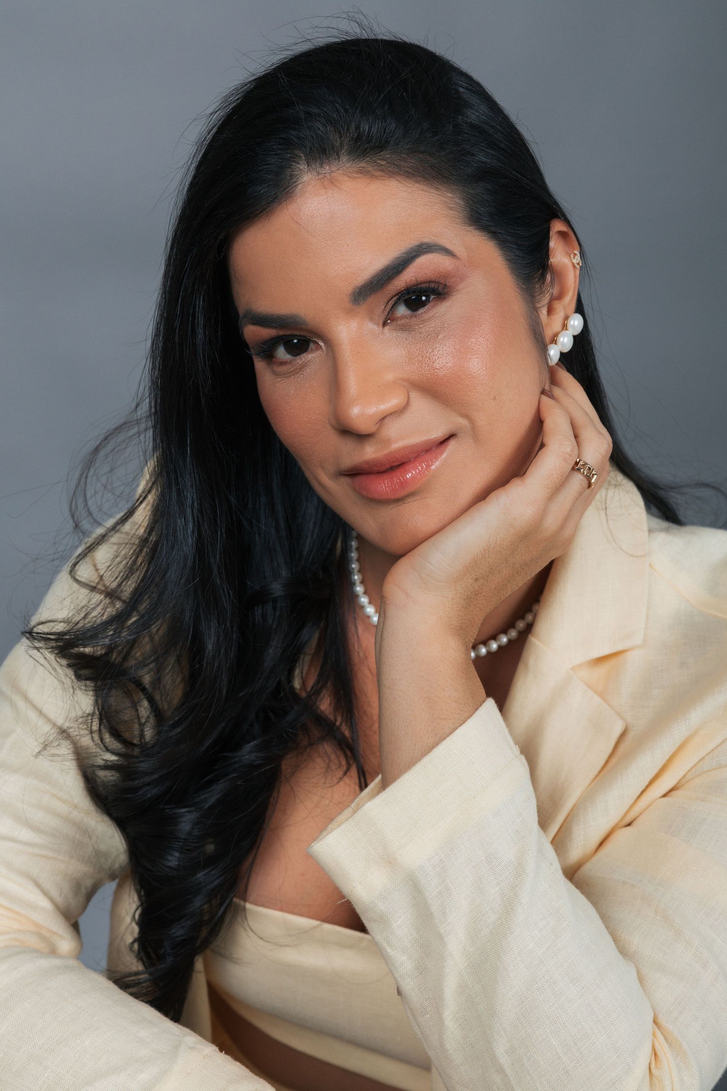 Professional portrait of a woman with long dark hair wearing a cream blazer and pearl accessories against a gray backdrop.