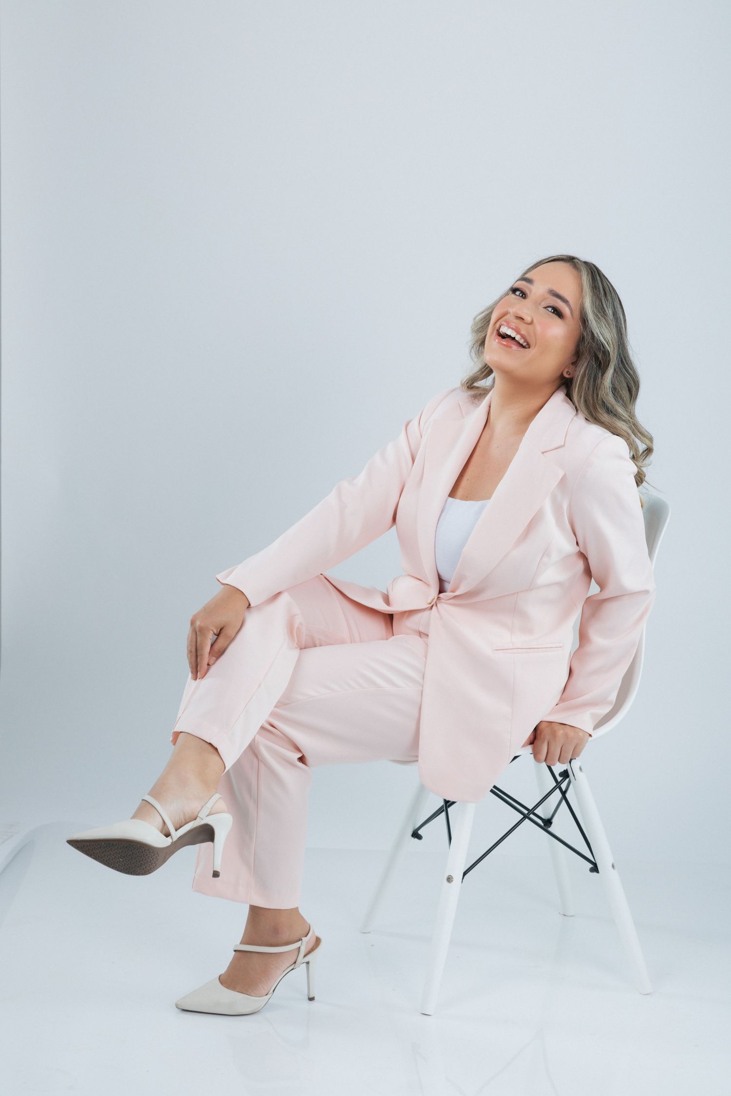 Someone wearing a blush pink business suit laughs joyfully while seated on a white chair against a gray background.