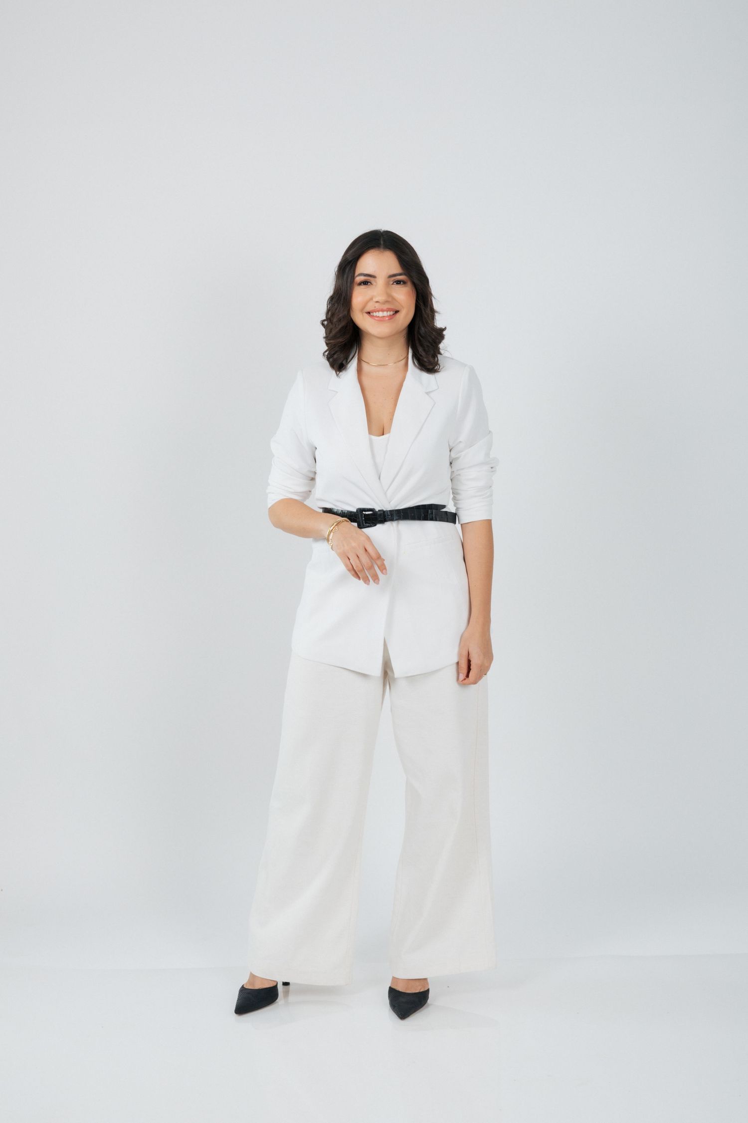 Stylish woman in white pantsuit with black belt standing confidently against gray backdrop.