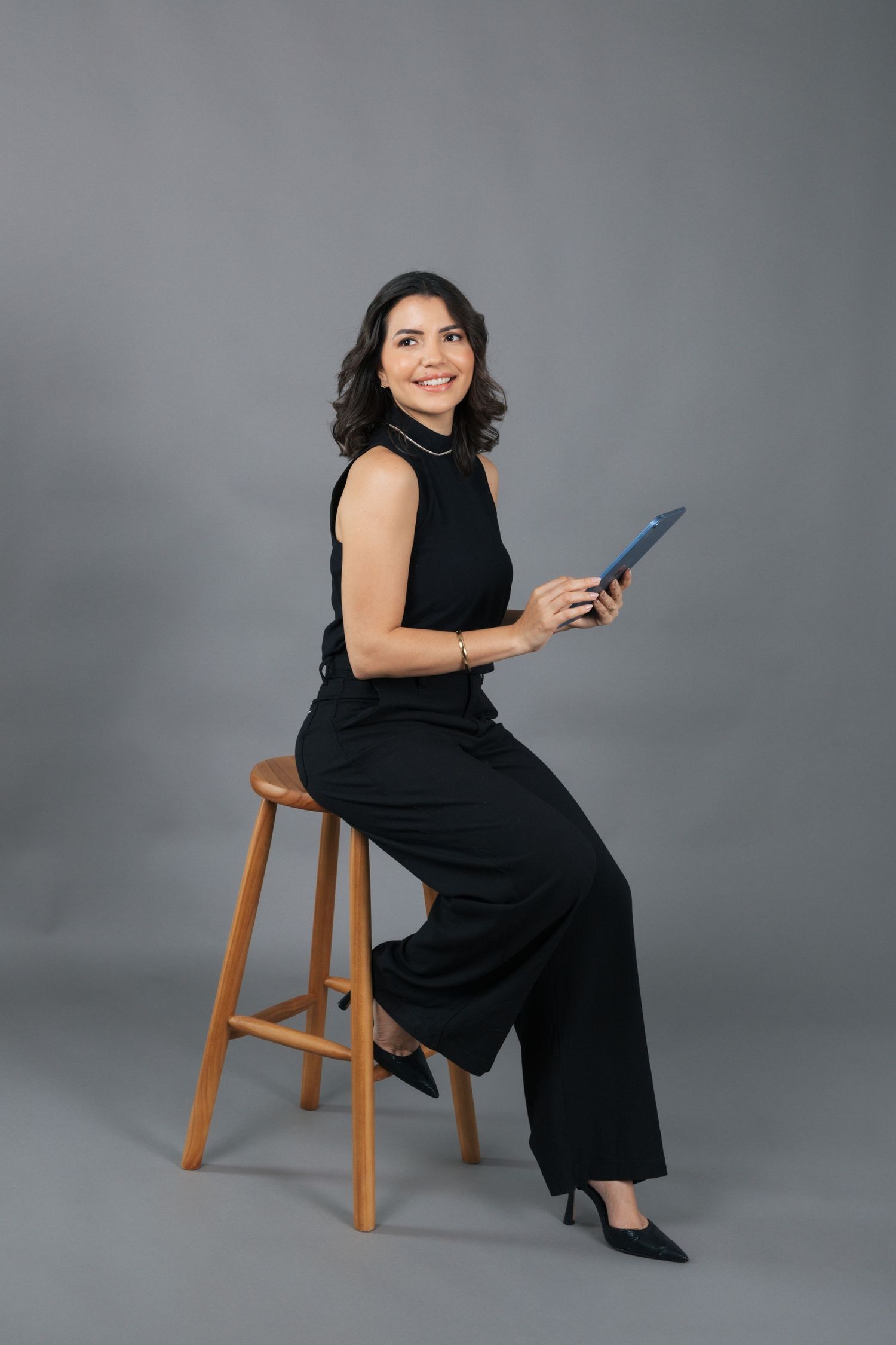 Professional seated on wooden stool holding tablet in all-black ensemble.
