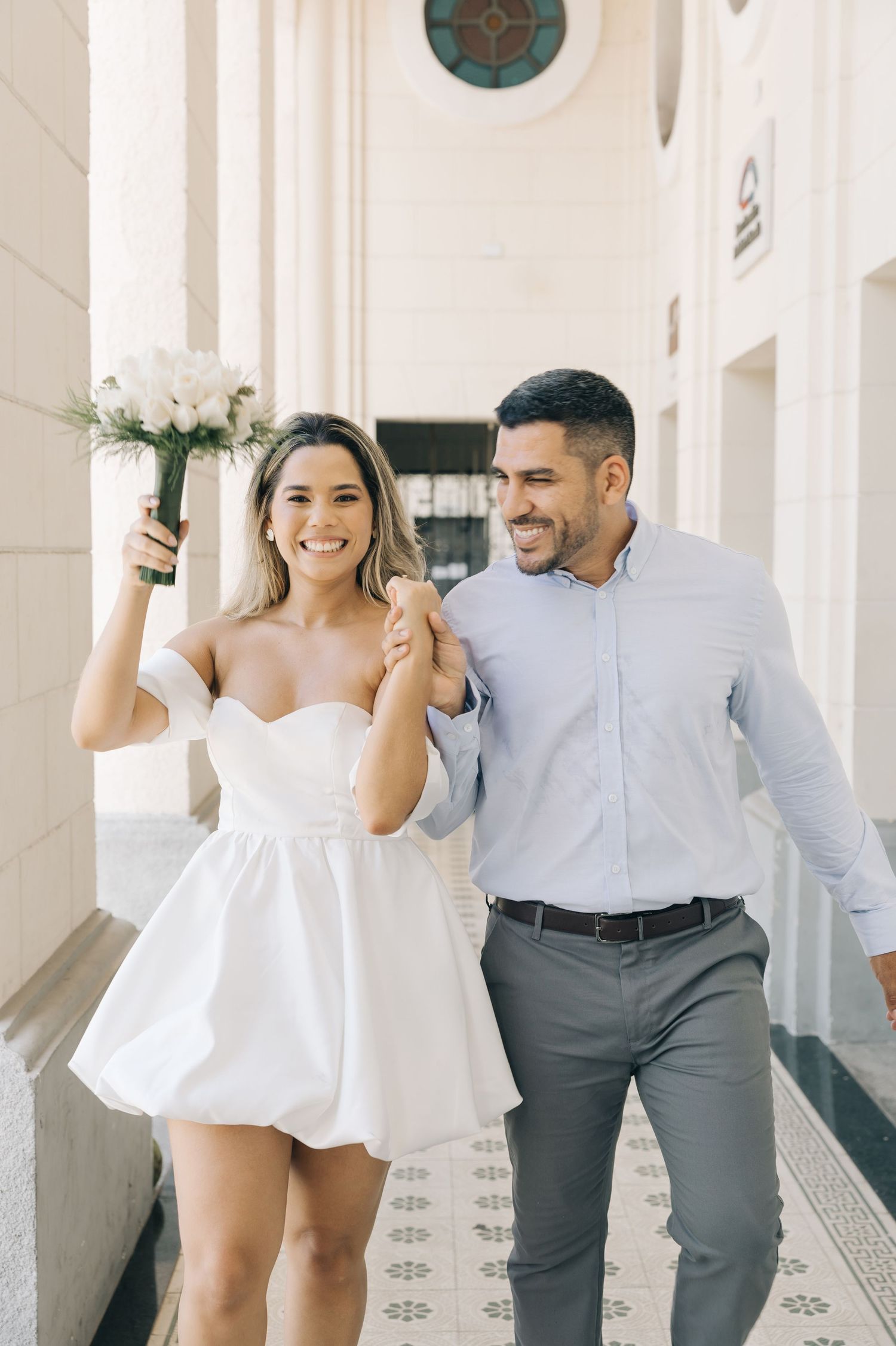 A joyful couple in wedding attire walking down an elegant corridor with white pillars and tiled flooring.