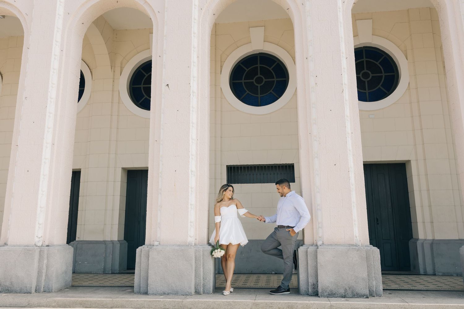 A couple stands together between elegant white stone columns with arched windows in a classic architectural setting.