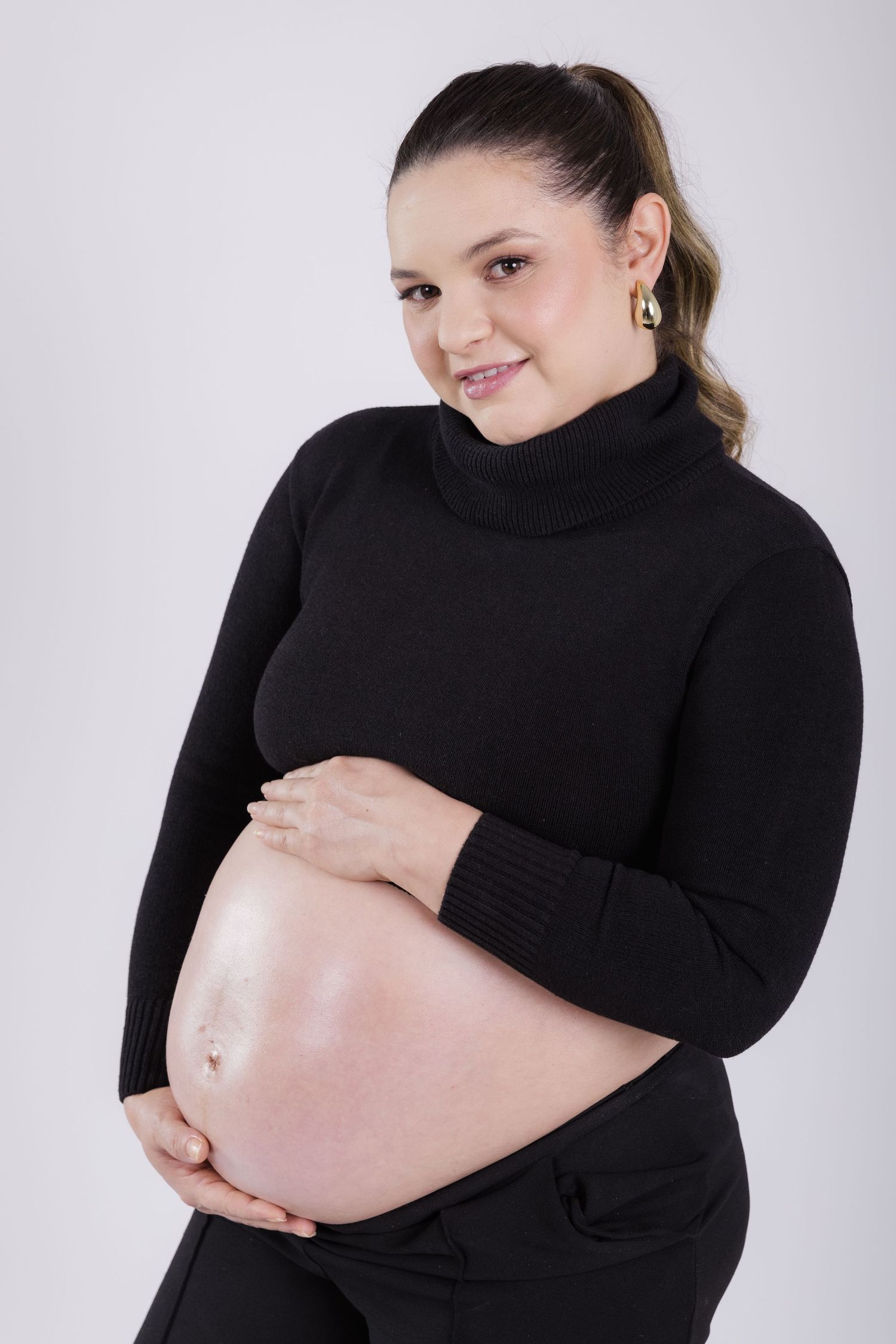Pregnant person wearing a black turtleneck sweater poses against light gray background while cradling their baby bump.