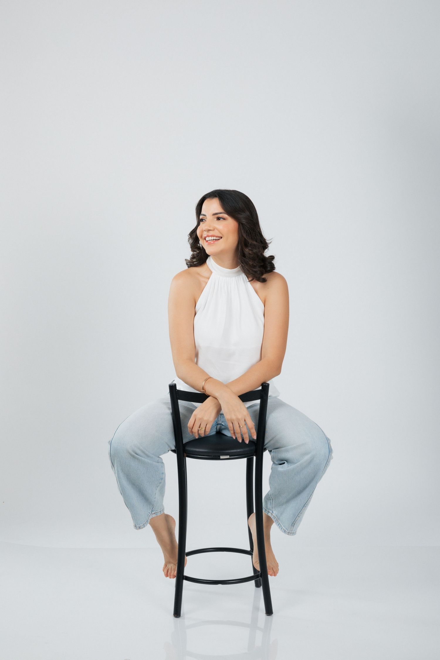 Casual studio pose on a black barstool wearing a white halter top and light denim jeans.