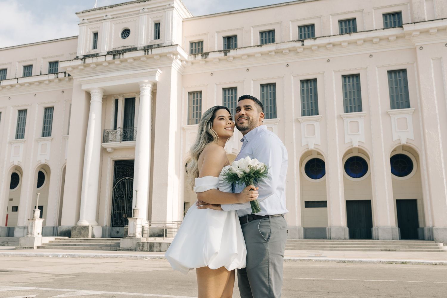 A pair pose together in wedding attire outside an elegant historic building with circular windows.