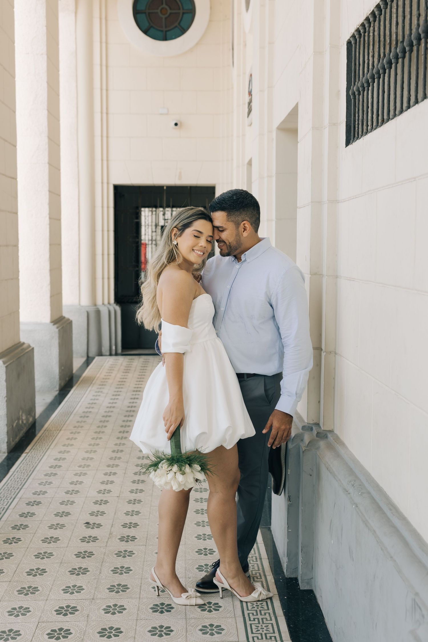 A romantic couple poses in an elegant corridor with ornate tiled floors and white walls.