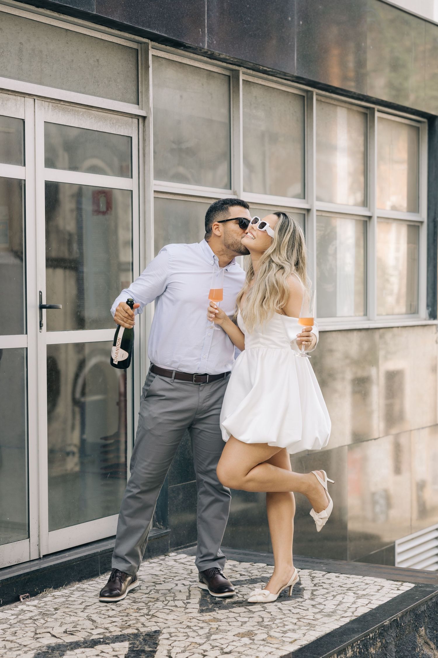 A playful couple poses together on a decorative sidewalk in front of a modern building.