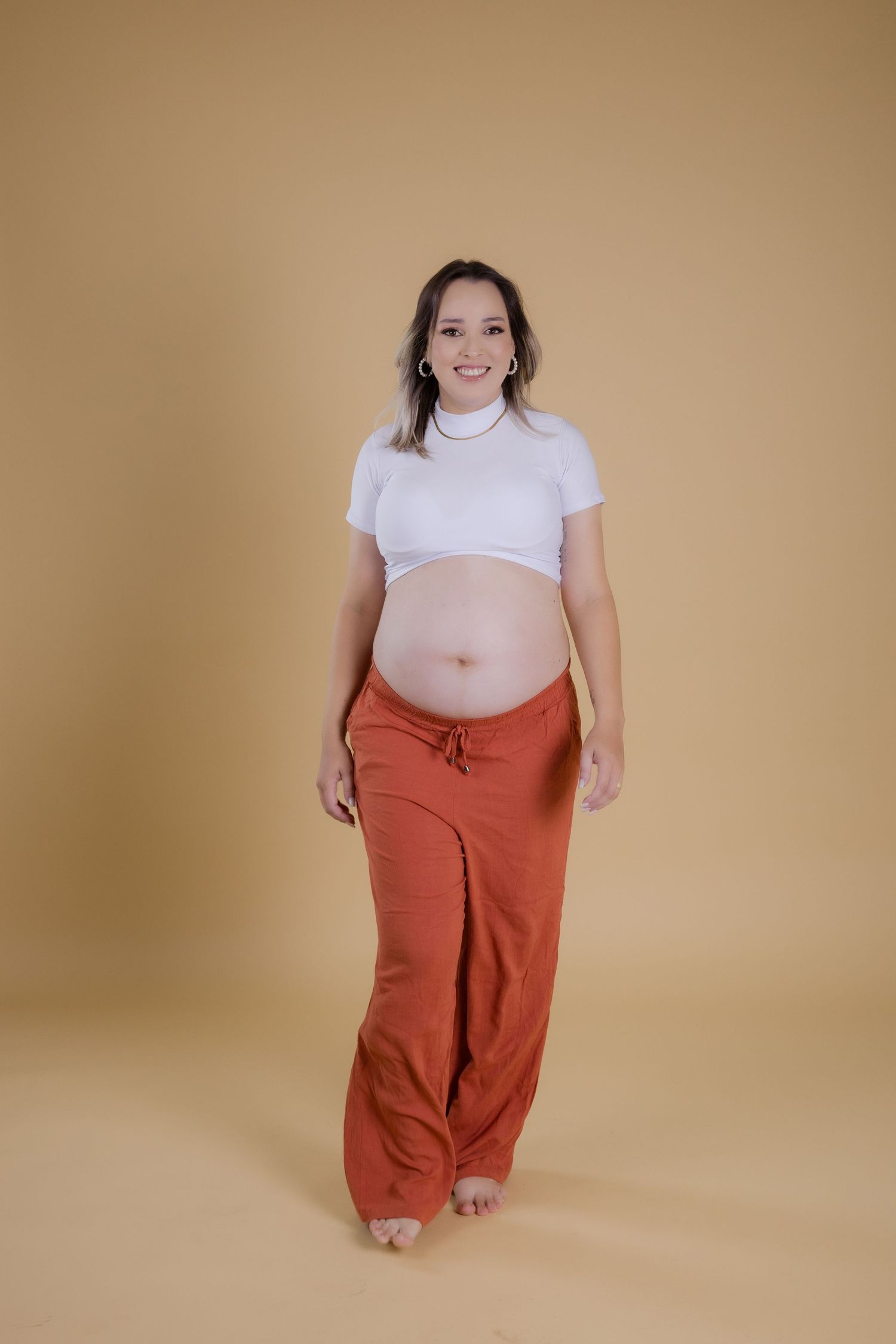 Pregnant woman wearing white turtleneck and orange pants stands barefoot against warm beige background.