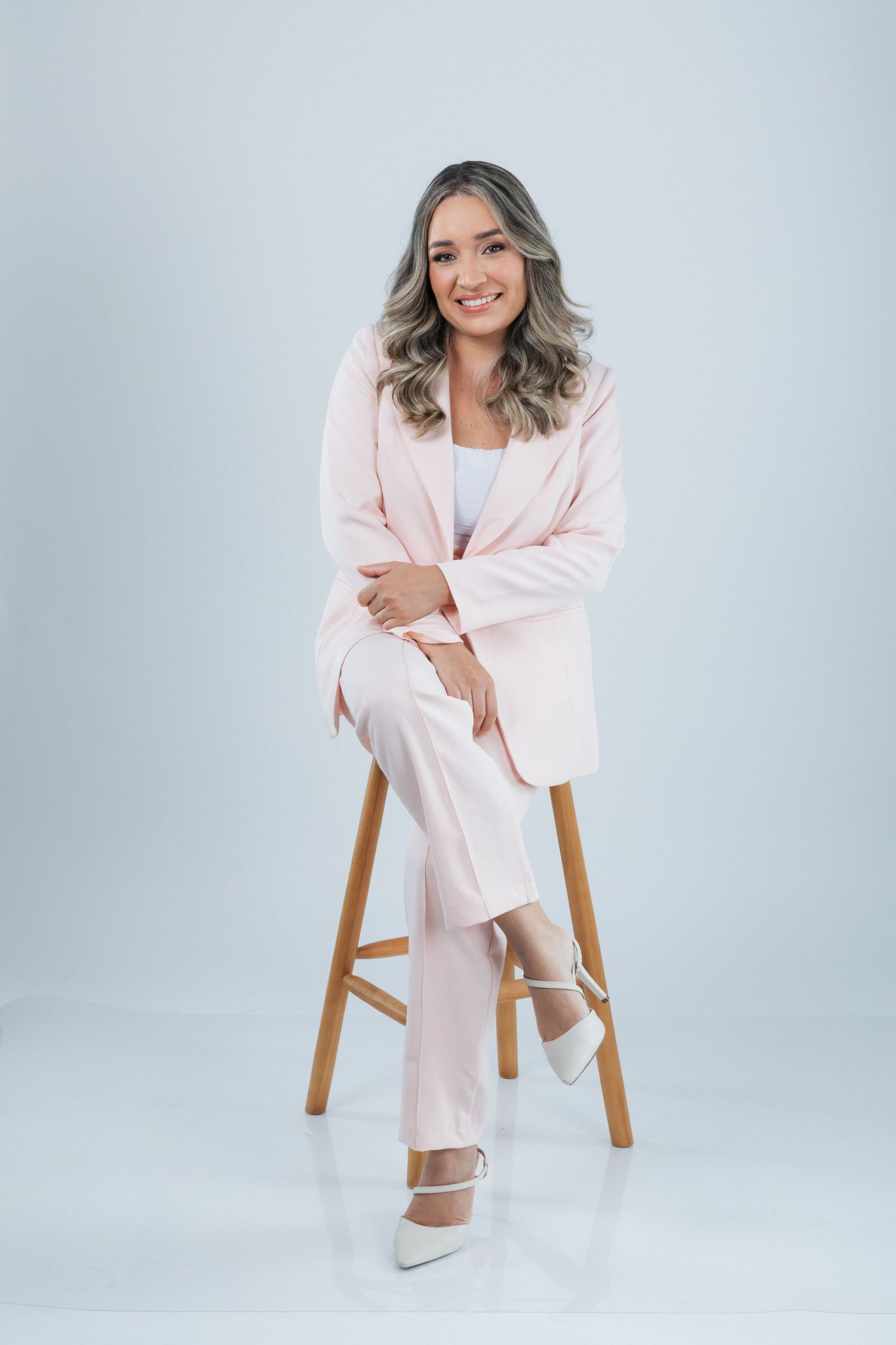 Person in a soft pink business suit and white heels sits elegantly on a wooden stool against a light gray background.