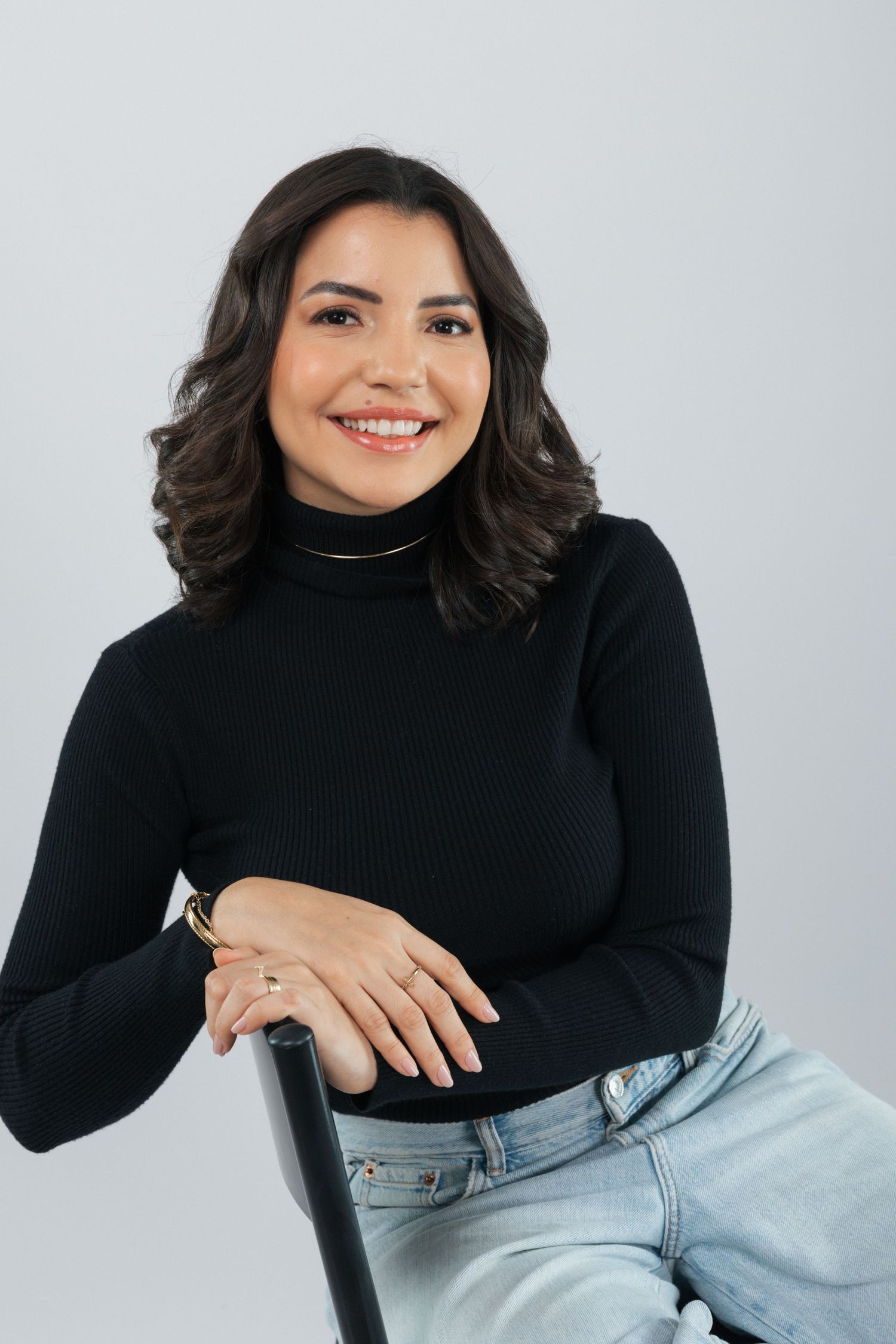 Smiling woman in black turtleneck sweater and light denim jeans sitting casually on a chair.