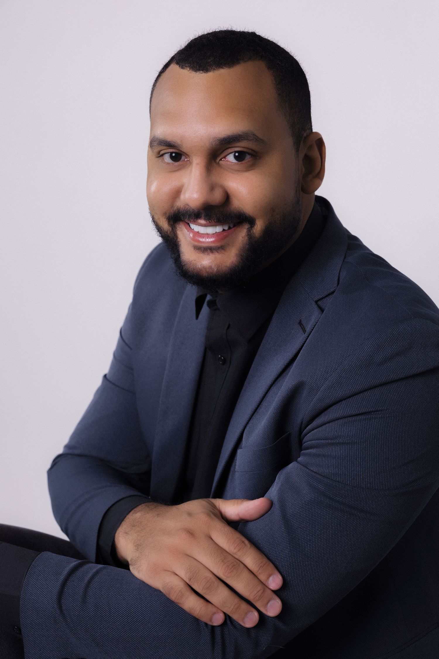 Professional headshot portrait of someone in a navy suit jacket smiling warmly against a light gray background.
