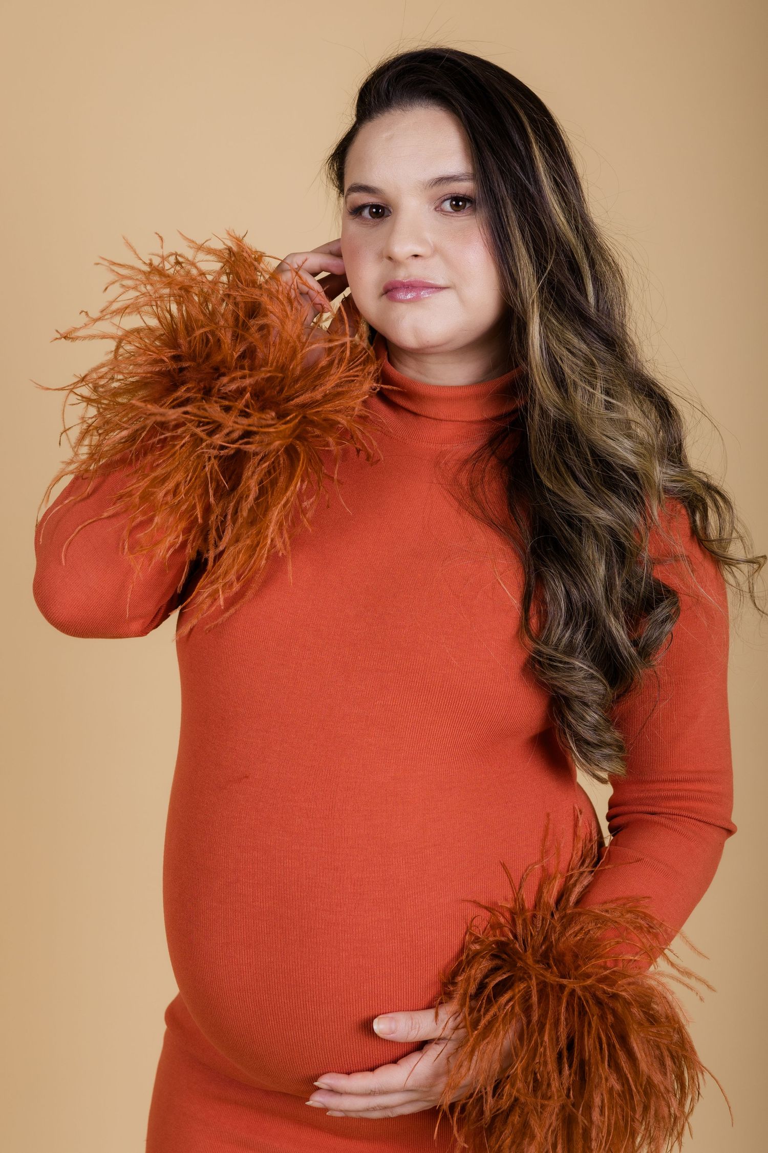 A person in a rust-colored turtleneck with long wavy hair poses with brown feather accessories against a beige backdrop.