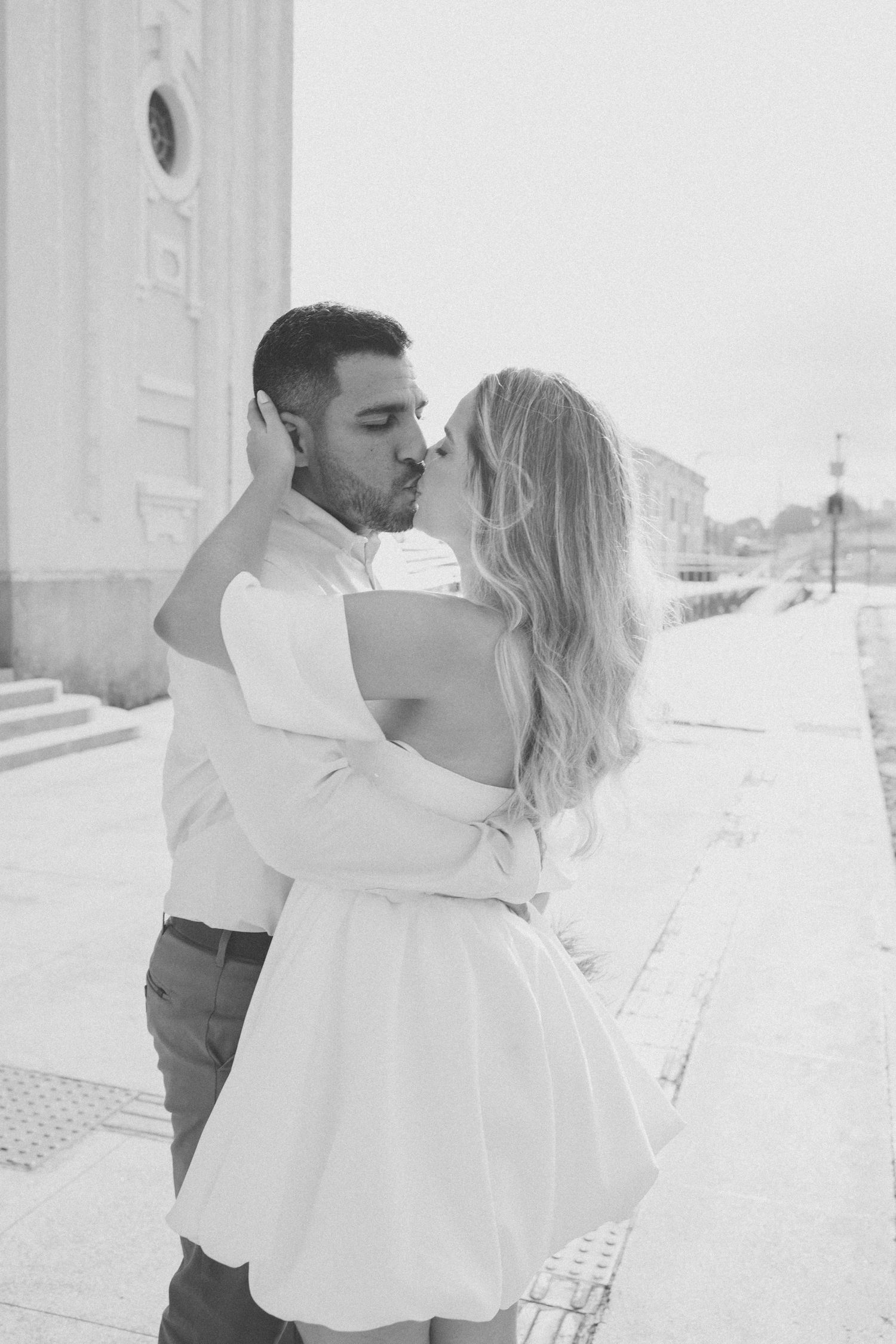 A romantic black and white photo of a couple embracing in front of a white building.