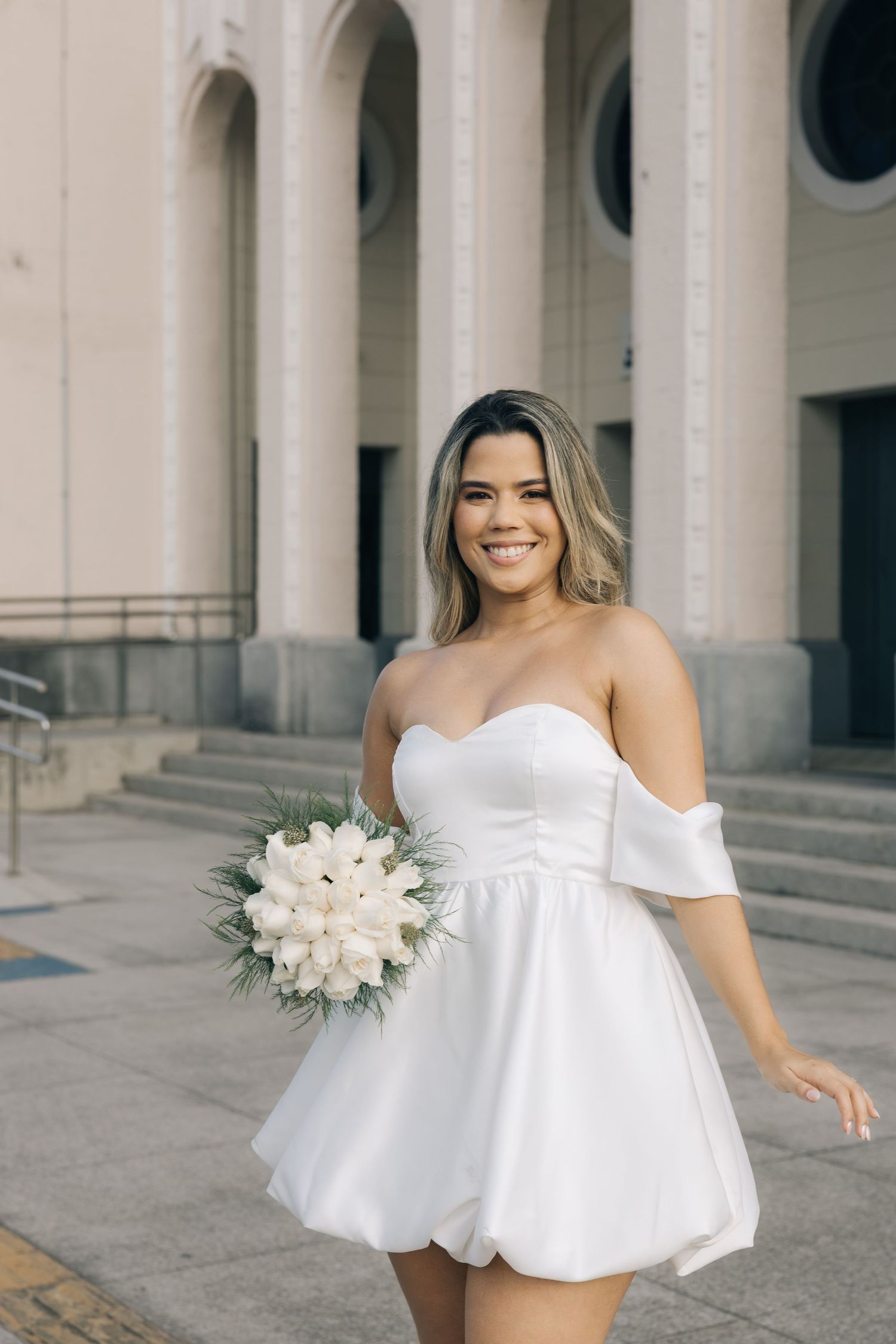 A bride in a short white off-shoulder wedding dress holds a white floral bouquet while standing on stone steps.