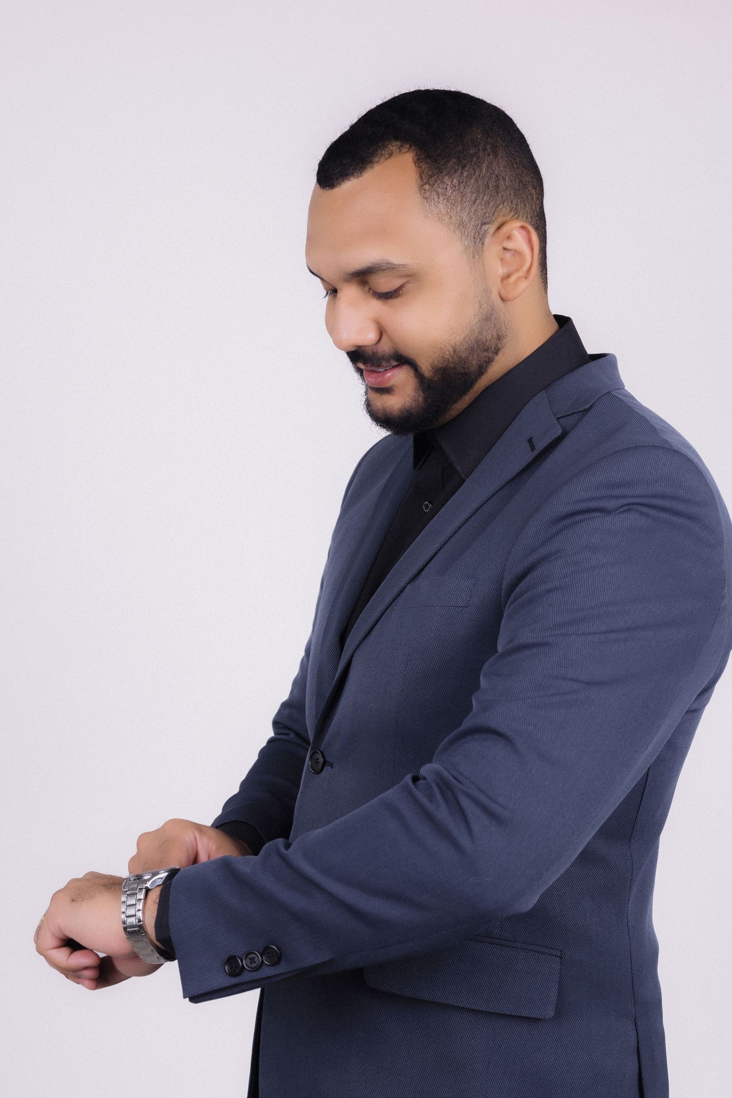 A person wearing a navy suit jacket and black shirt checks their wrist watch against a light background.