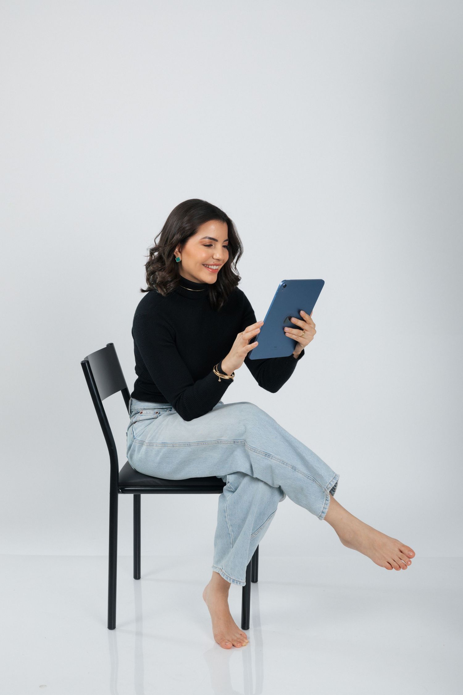 Person sitting barefoot on chair reads from blue tablet while wearing black top and light denim.