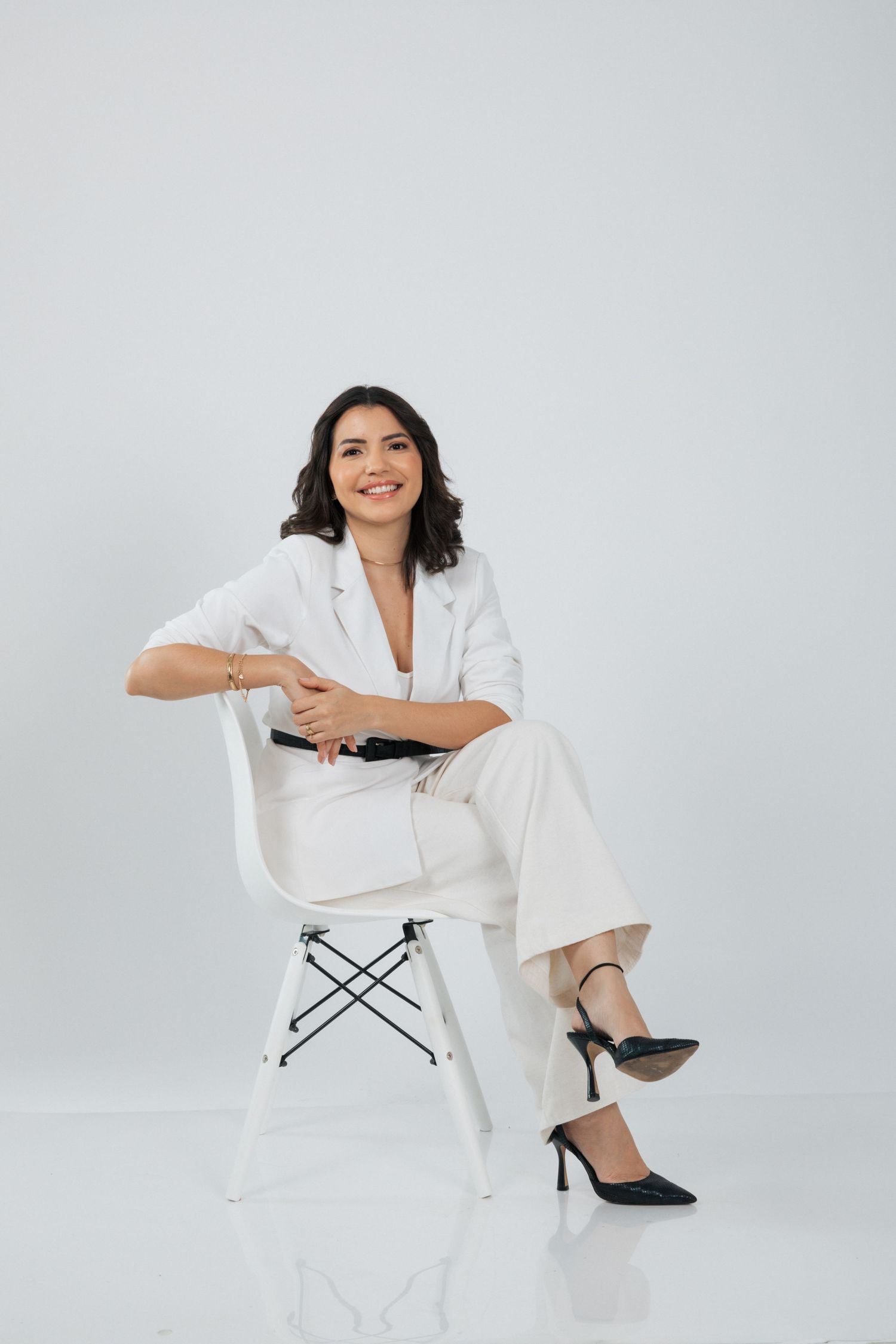 Stylish model in white business suit seated elegantly on modern white chair against neutral backdrop.