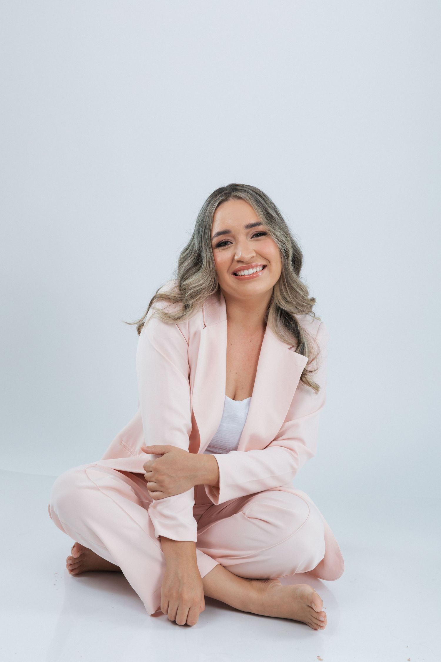Person in pink business suit sitting cross-legged on white background with casual, friendly expression.
