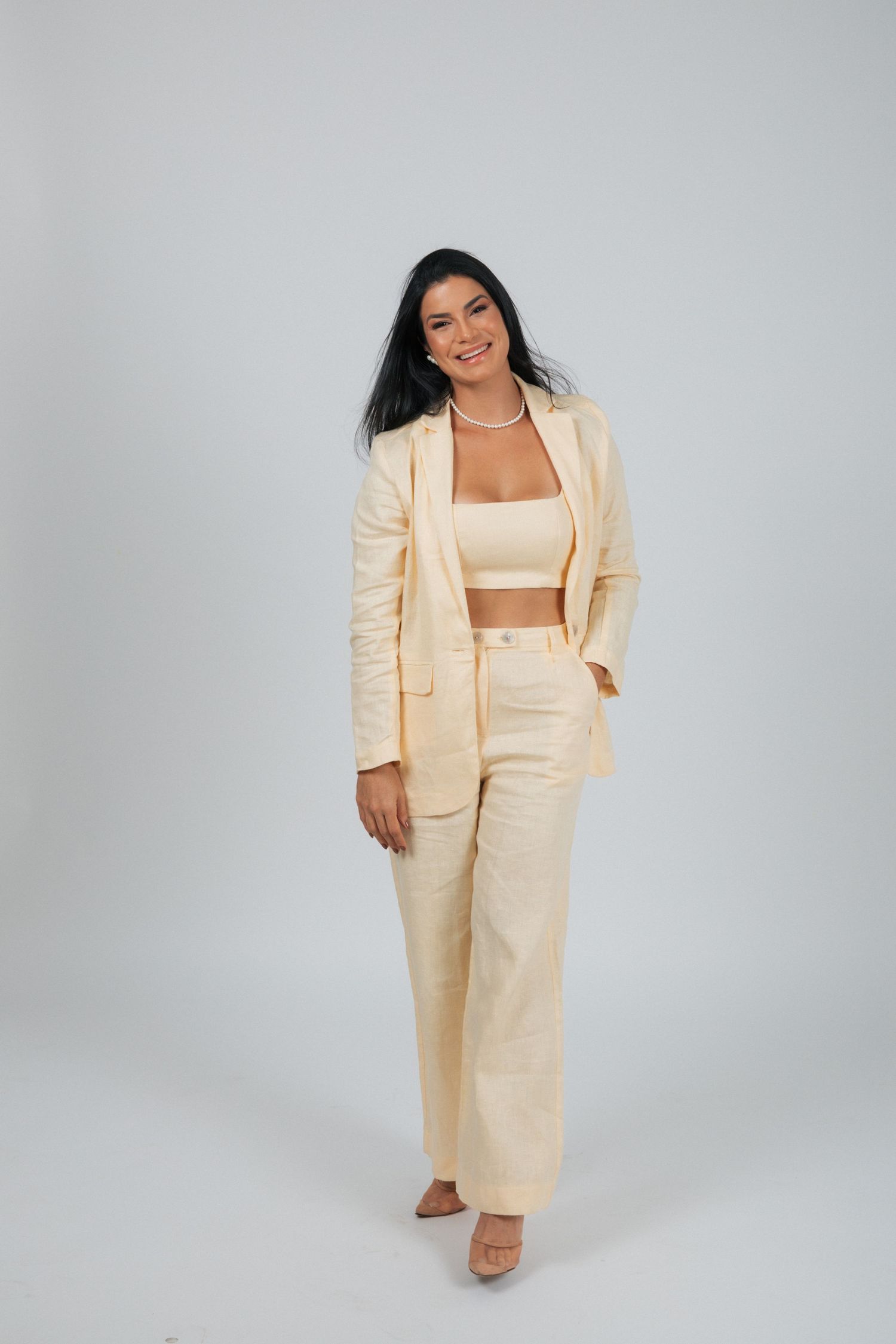 A person wearing a cream colored pantsuit with a crop top stands confidently against a gray background.