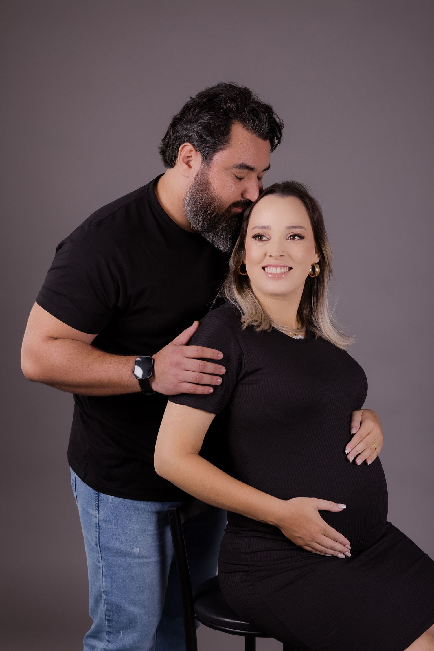 A pregnant couple in matching black outfits pose for a maternity photo against a gray studio backdrop.